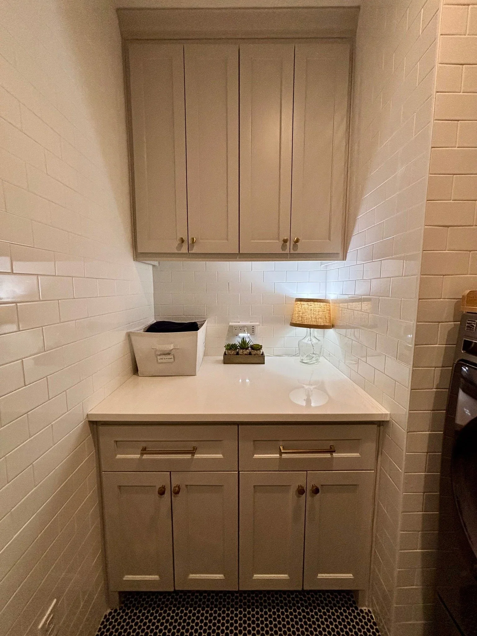 A tidy laundry folding station by A.T. Home Organizing with grey cabinets, white subway tile, and a small lamp.