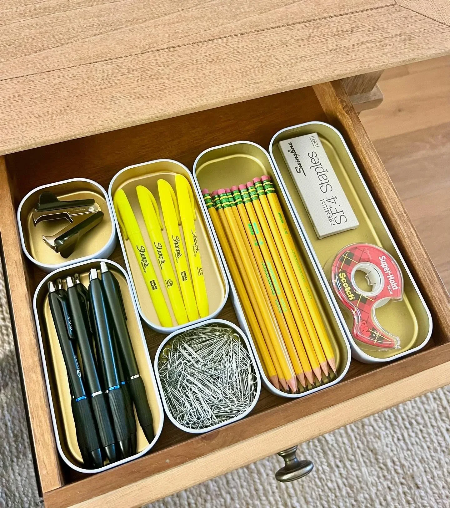 Organized desk drawer with office supplies in white dividers, including pens, pencils, and Sharpies.