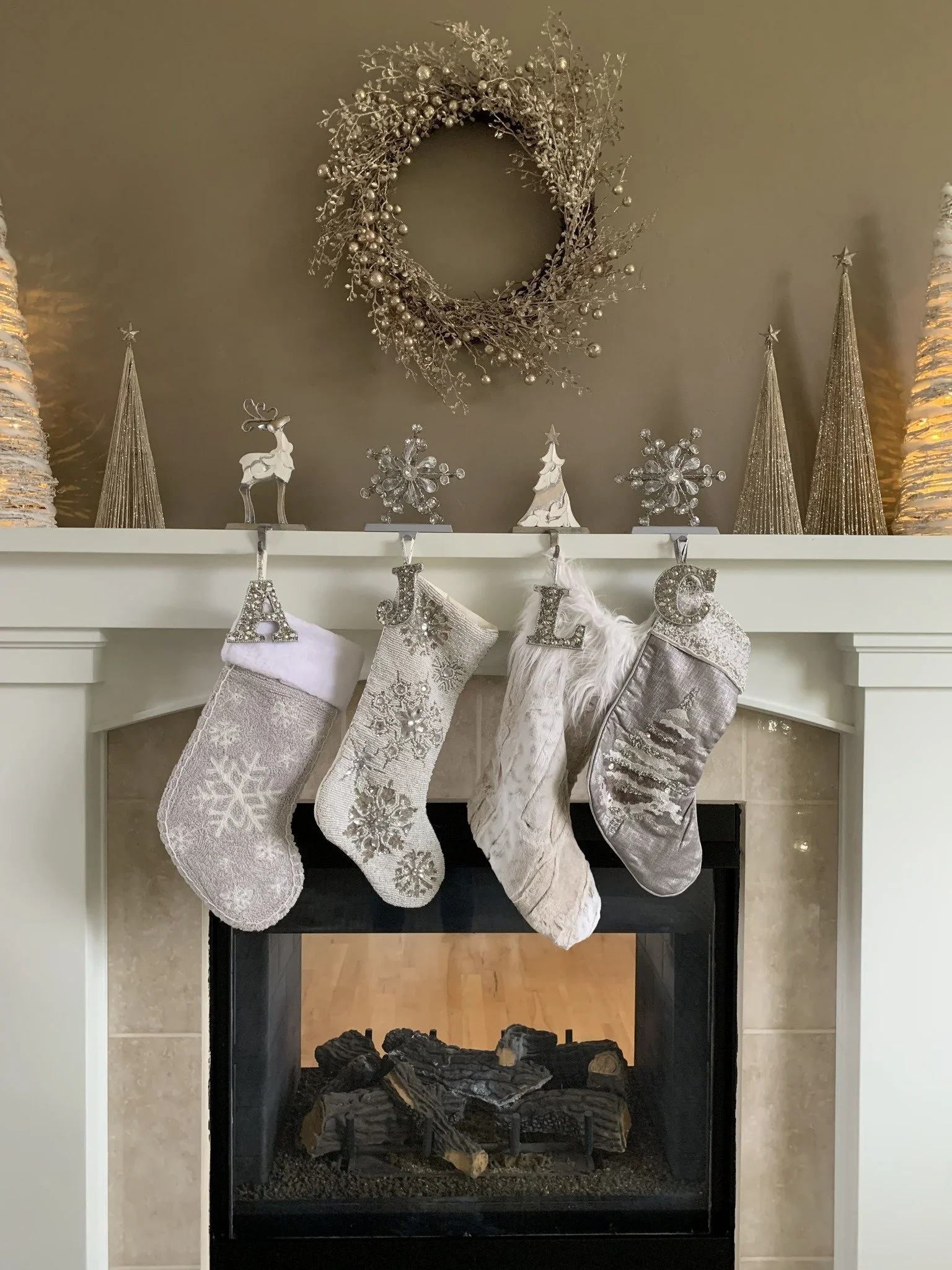 A fireplace mantel decorated with silver and white Christmas stockings, a gold wreath, and glittery trees.