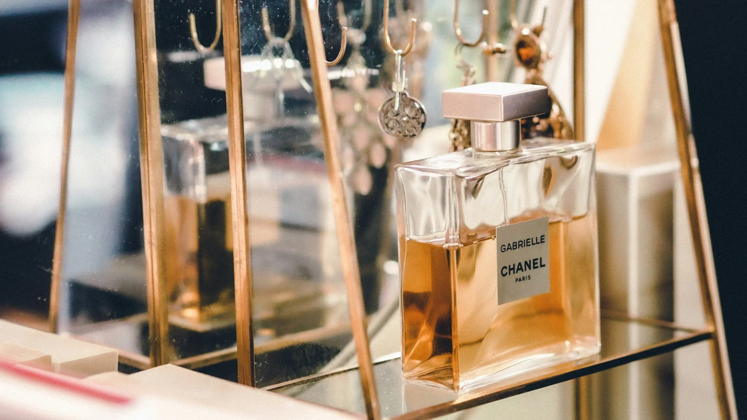 Organized glass vanity shelf by Ethereal Edits featuring a Gabrielle Chanel perfume bottle and a gold jewelry stand.
