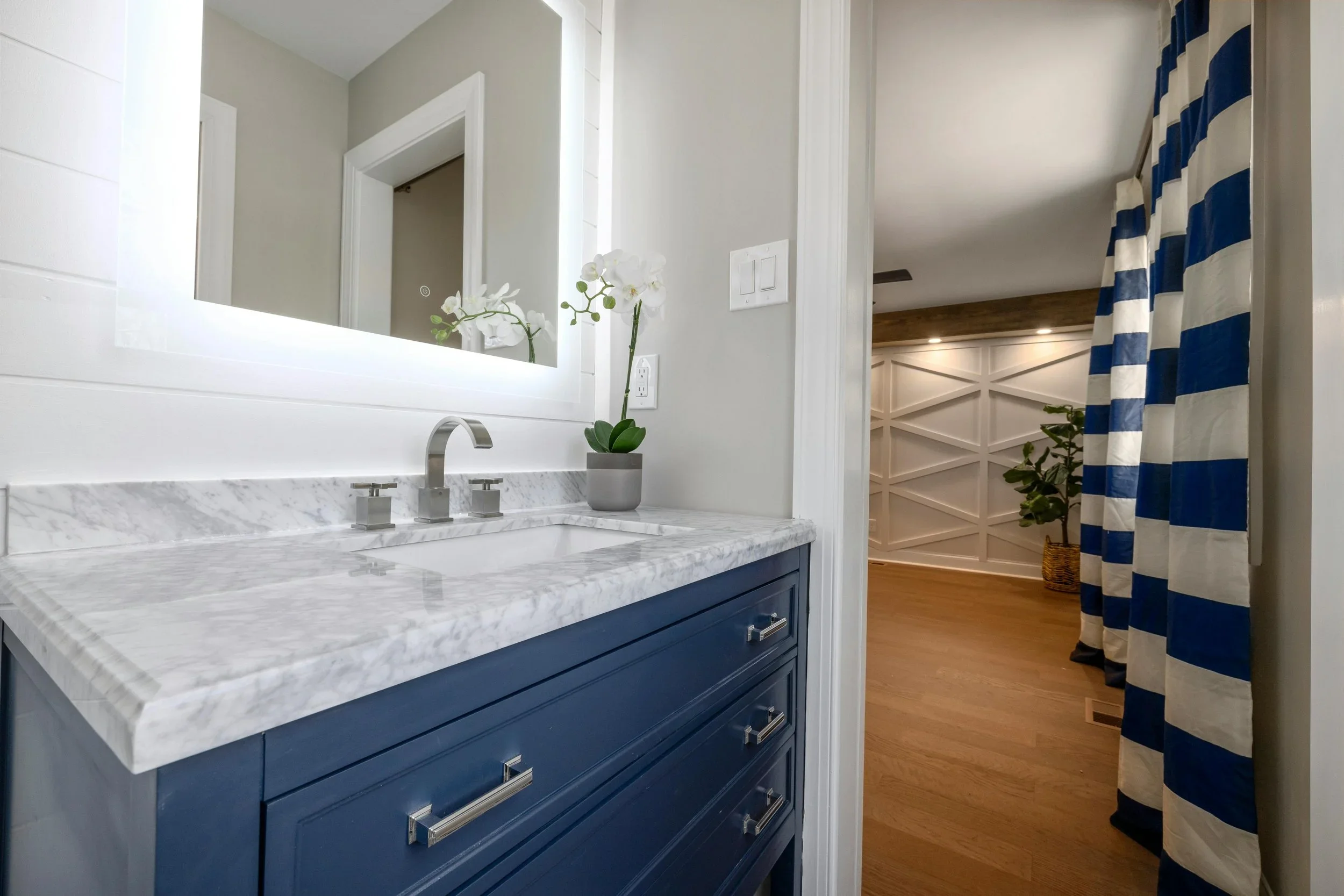 A modern bathroom organized by The Tidy Girl in MetroWest, featuring a navy blue vanity with a marble top and an illuminated mirror next to nautical striped curtains.