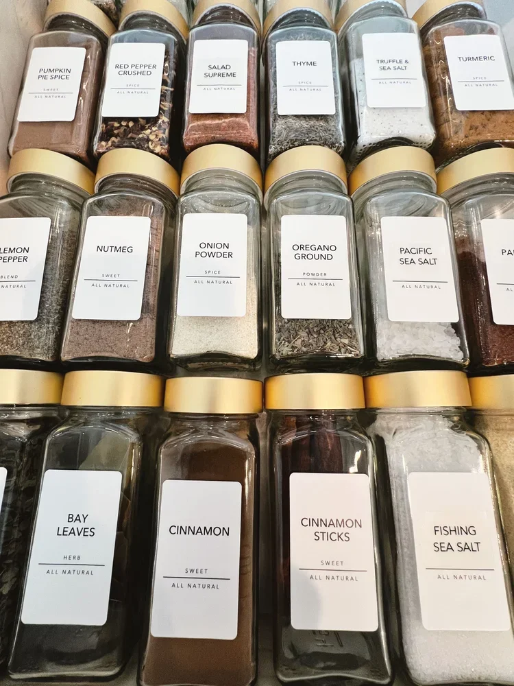 Uniformly labeled glass spice jars with gold lids in a neatly organized drawer.