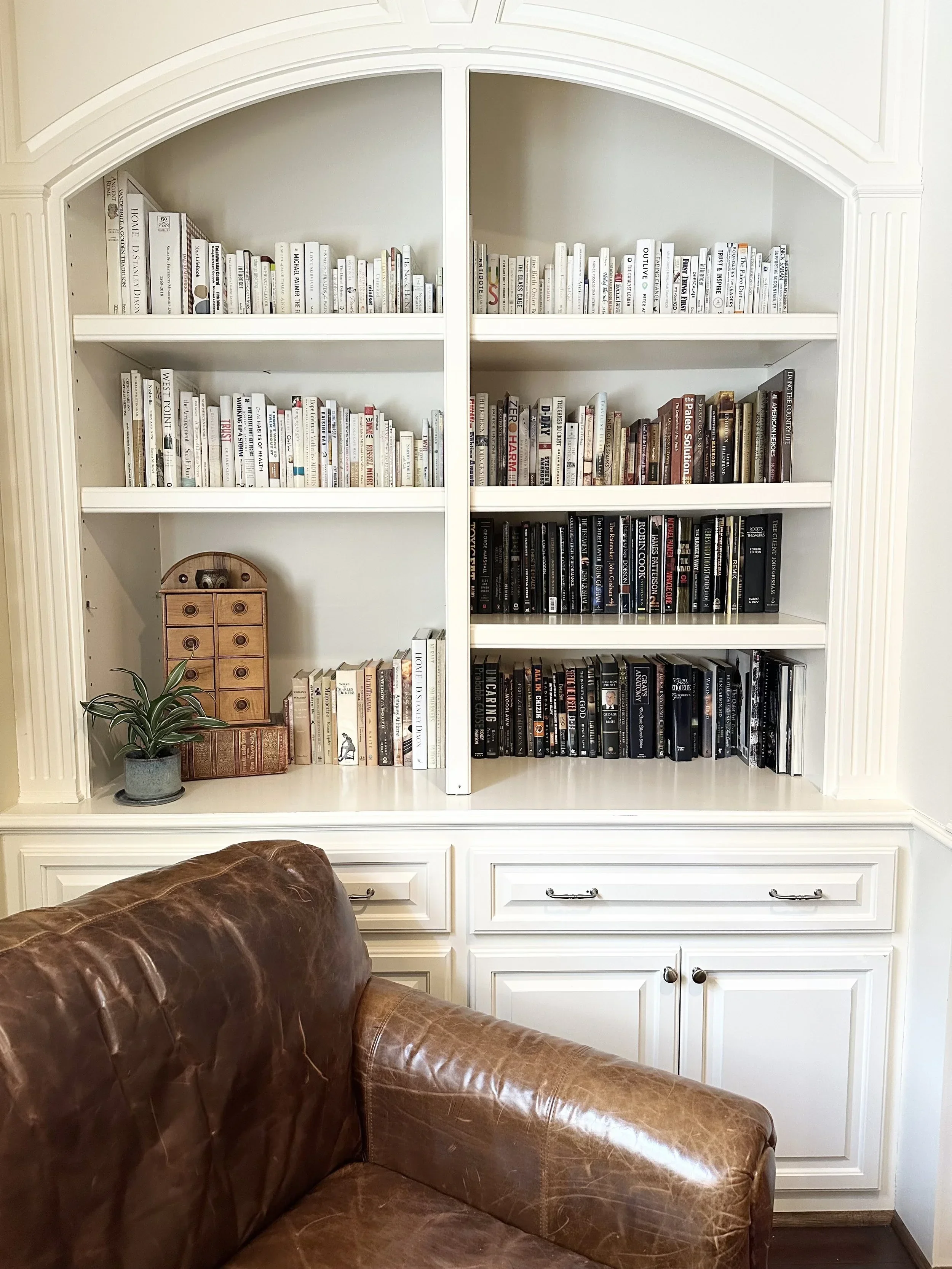 Neatly organized white built-in bookshelf by Refined Living Co. in Nashville, featuring a brown leather armchair.