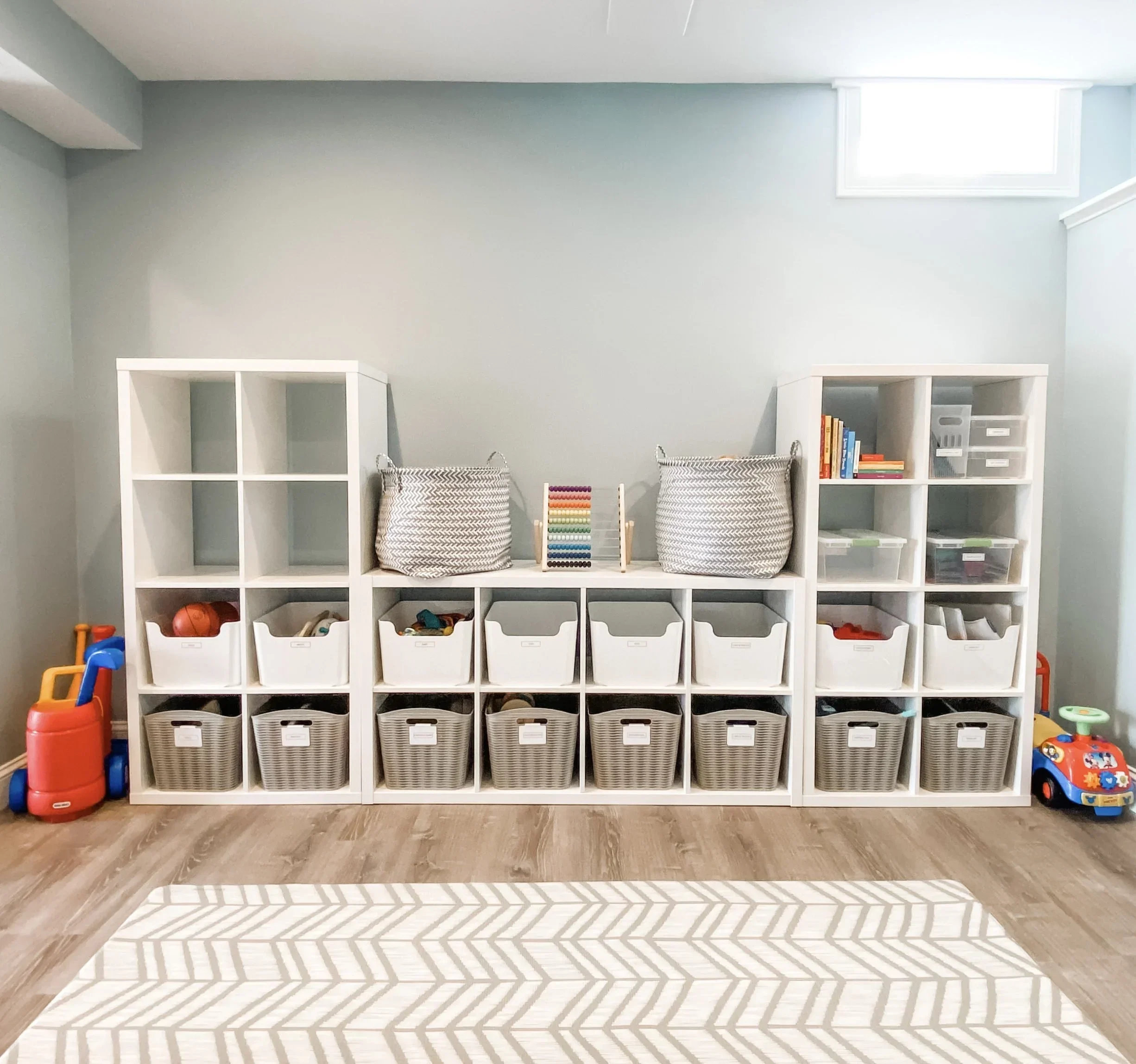 Organized white cube storage unit in a playroom with labeled grey and white bins.