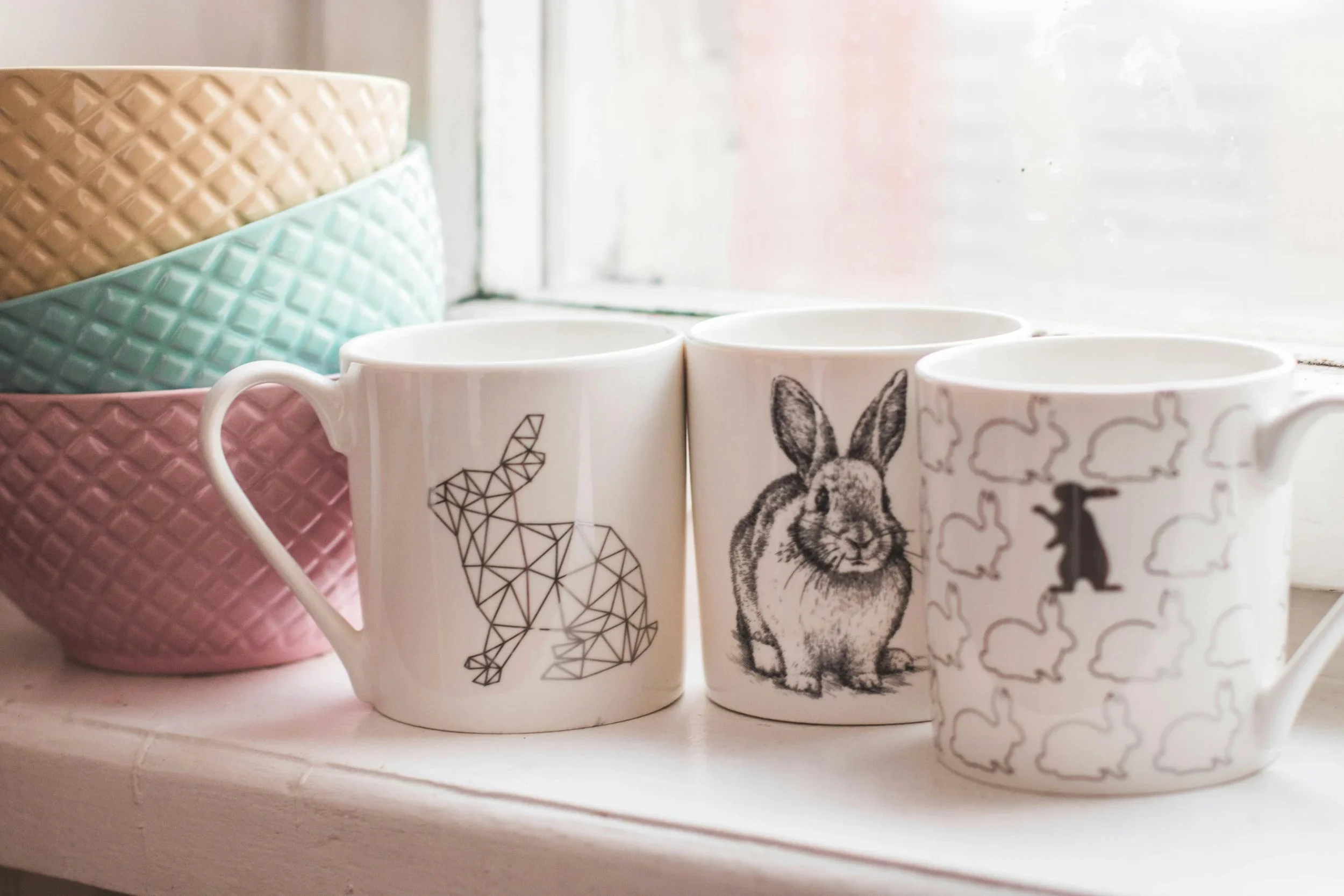 Three rabbit-themed mugs and a stack of textured pastel bowls on an organized white counter.