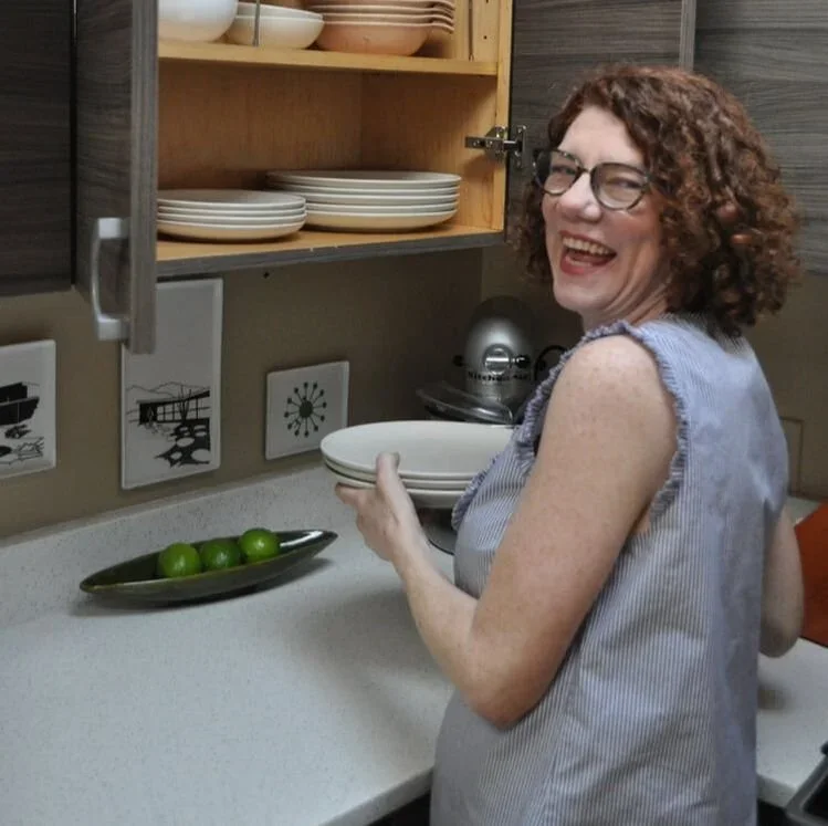 Professional organizer Jane Corkery Hahn laughing while organizing plates in a modern kitchen cabinet.
