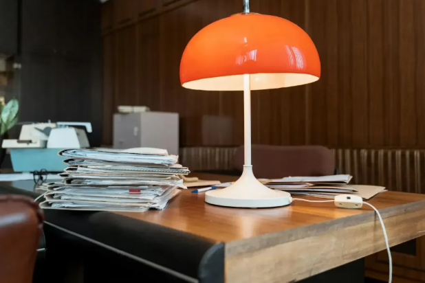 A cluttered wooden office desk with a large stack of papers and a bright orange retro mushroom lamp.