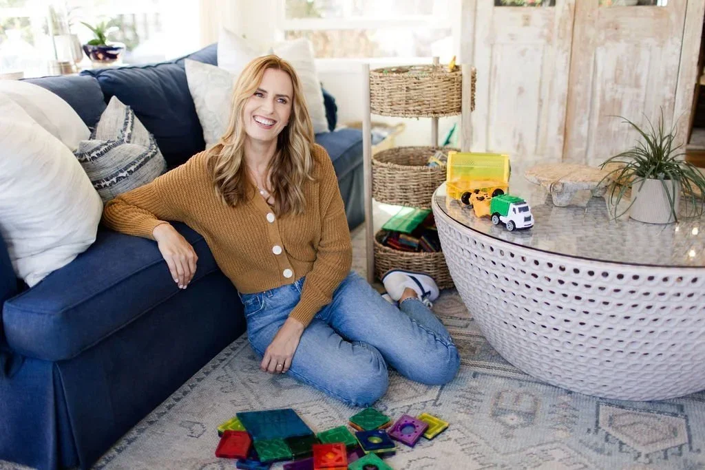 Professional organizer from The Organized Mommy Co. sitting in an organized living room with toys and woven storage baskets.