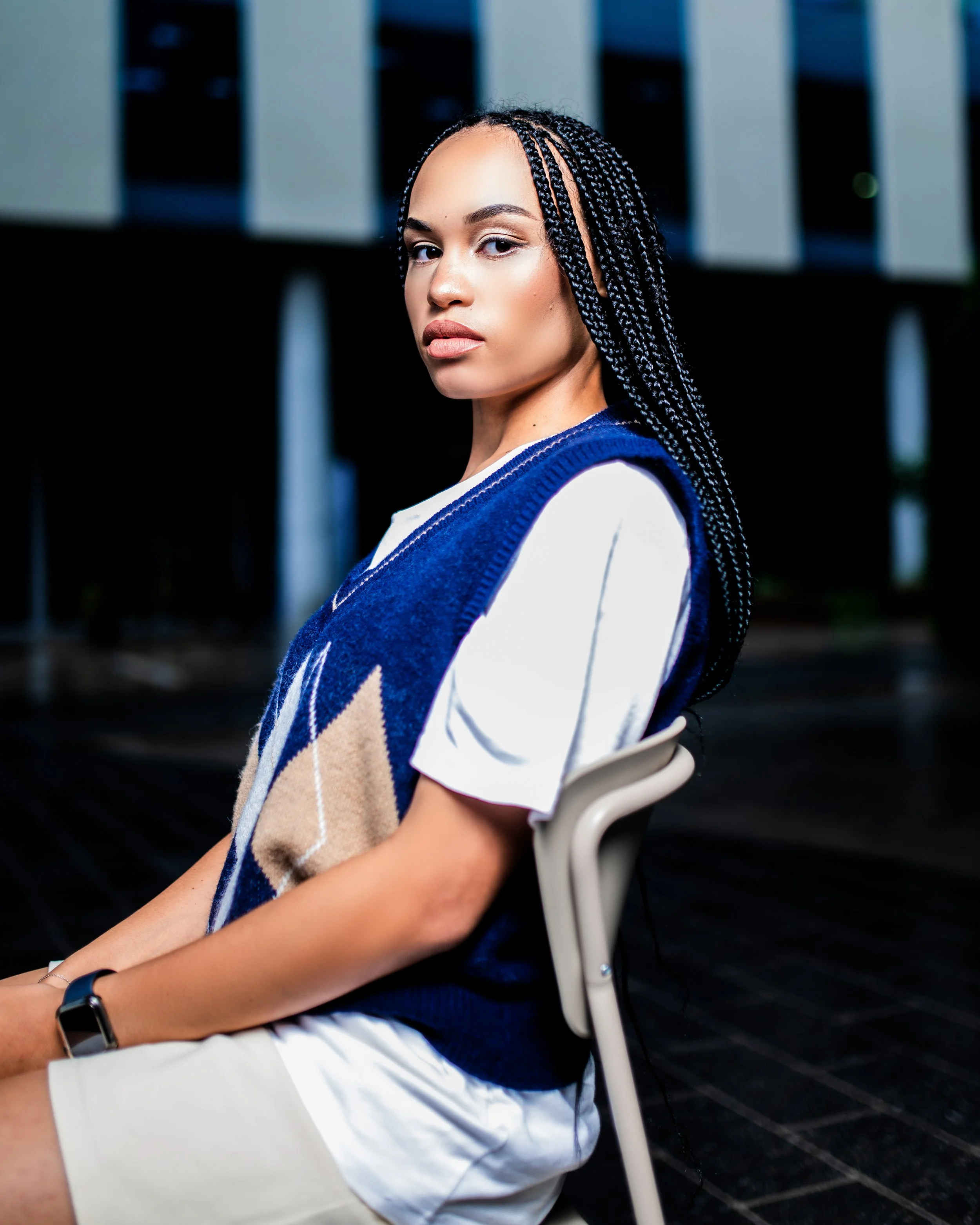 Person seated on a chair, wearing a blue and beige vest over a white shirt, with braided hair, in an outdoor setting.  Photographed in Rosebank, Johannesburg. 