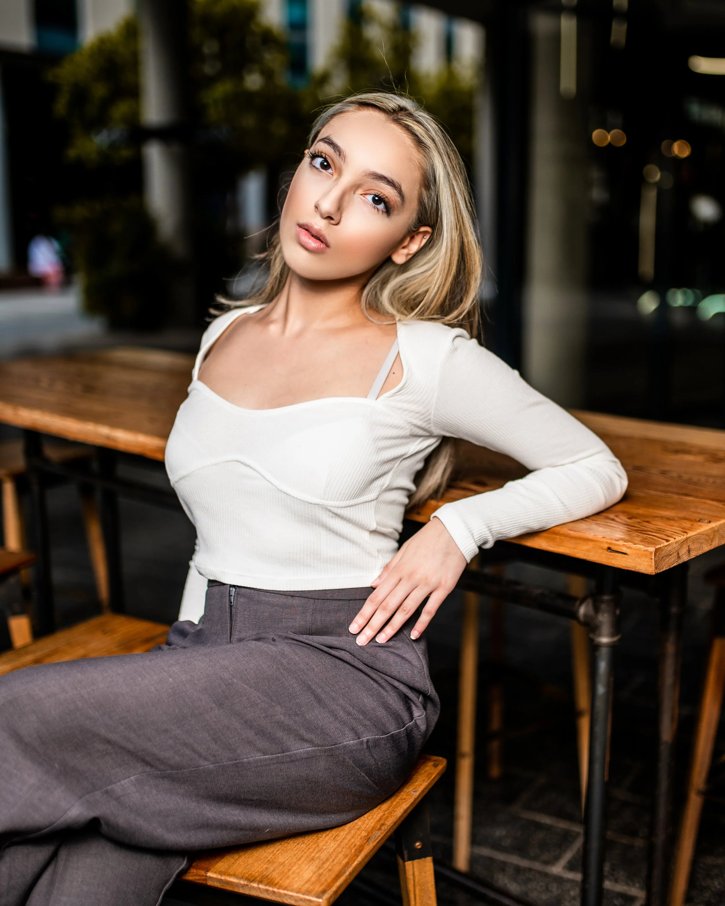 Person sitting on a wooden bench, leaning back against a table, wearing a white long-sleeve top and high-waisted gray pants.  Photographed in Rosebank, Johannesburg. 