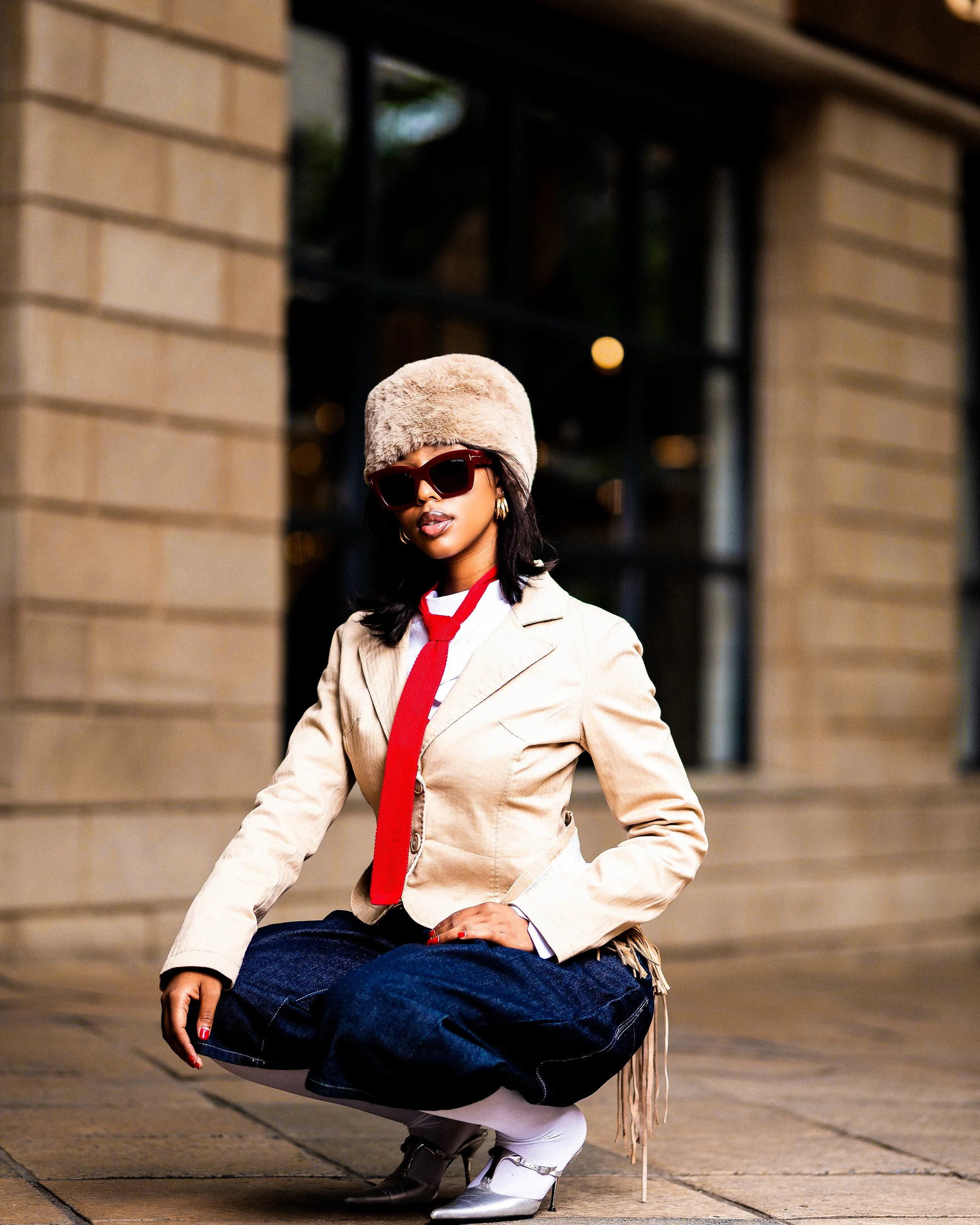 Person wearing a beige jacket, red tie, denim trousers, furry hat, and sunglasses, crouching on a city street. Photographed in Melrose Arch, South Africa.
