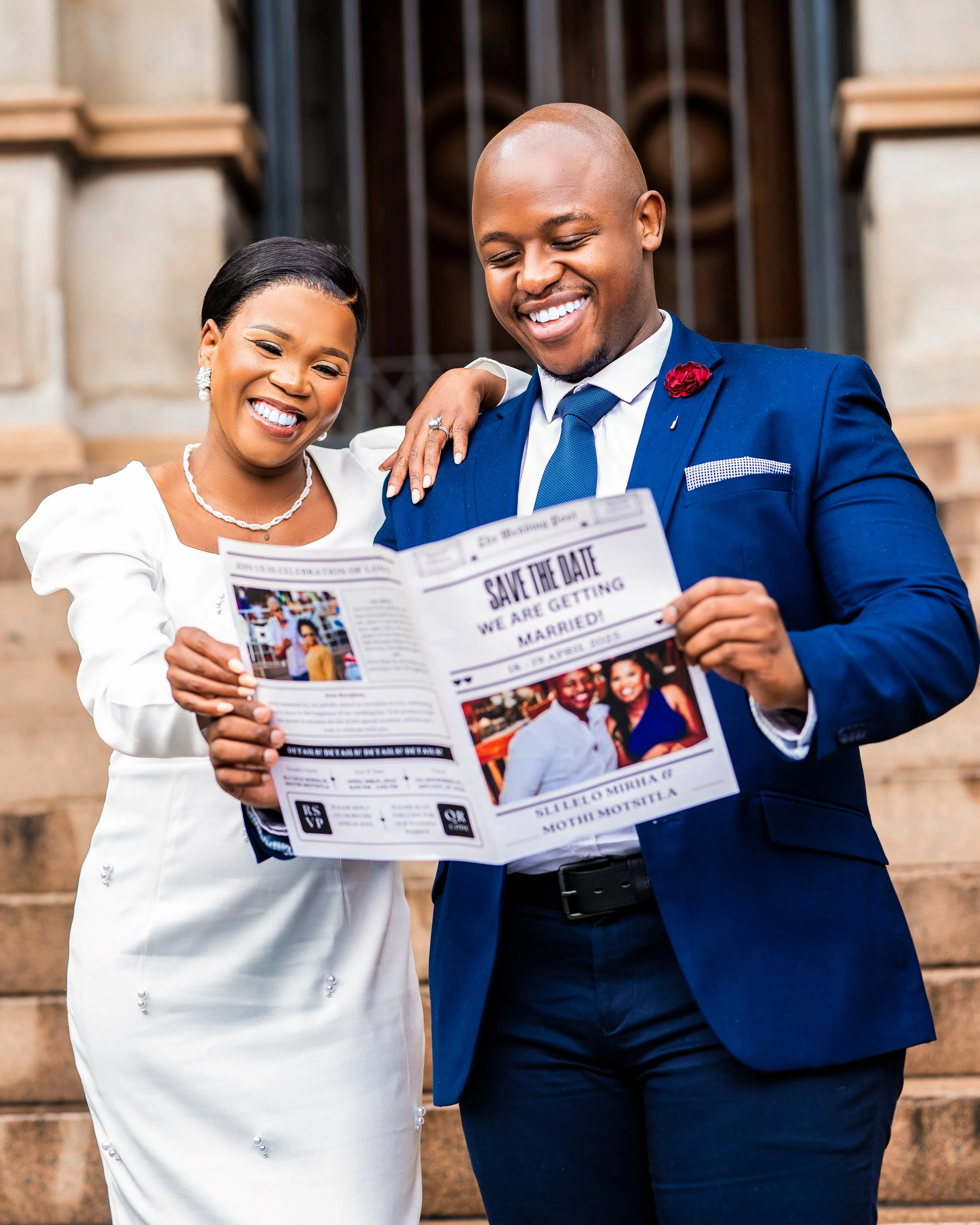 A smiling couple dressed formally holding a "Save the Date" wedding announcement newspaper on outdoor steps. Photographed in Pretoria, South Africa. 