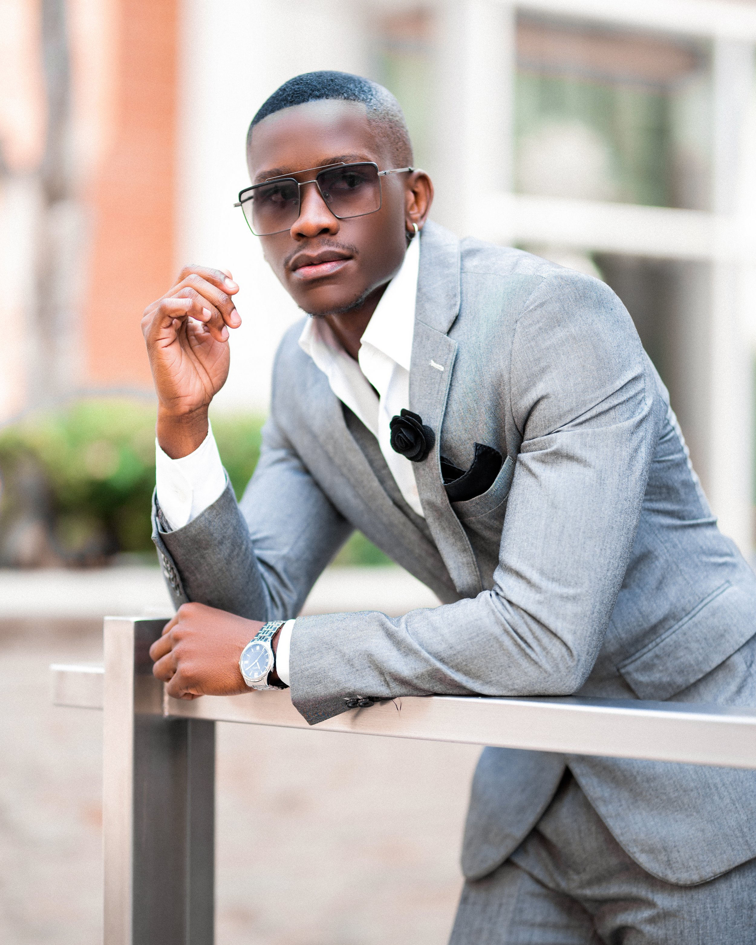 Man in a gray suit with sunglasses leaning on a railing outdoors. Photographed in Melrose, Johannesburg. 