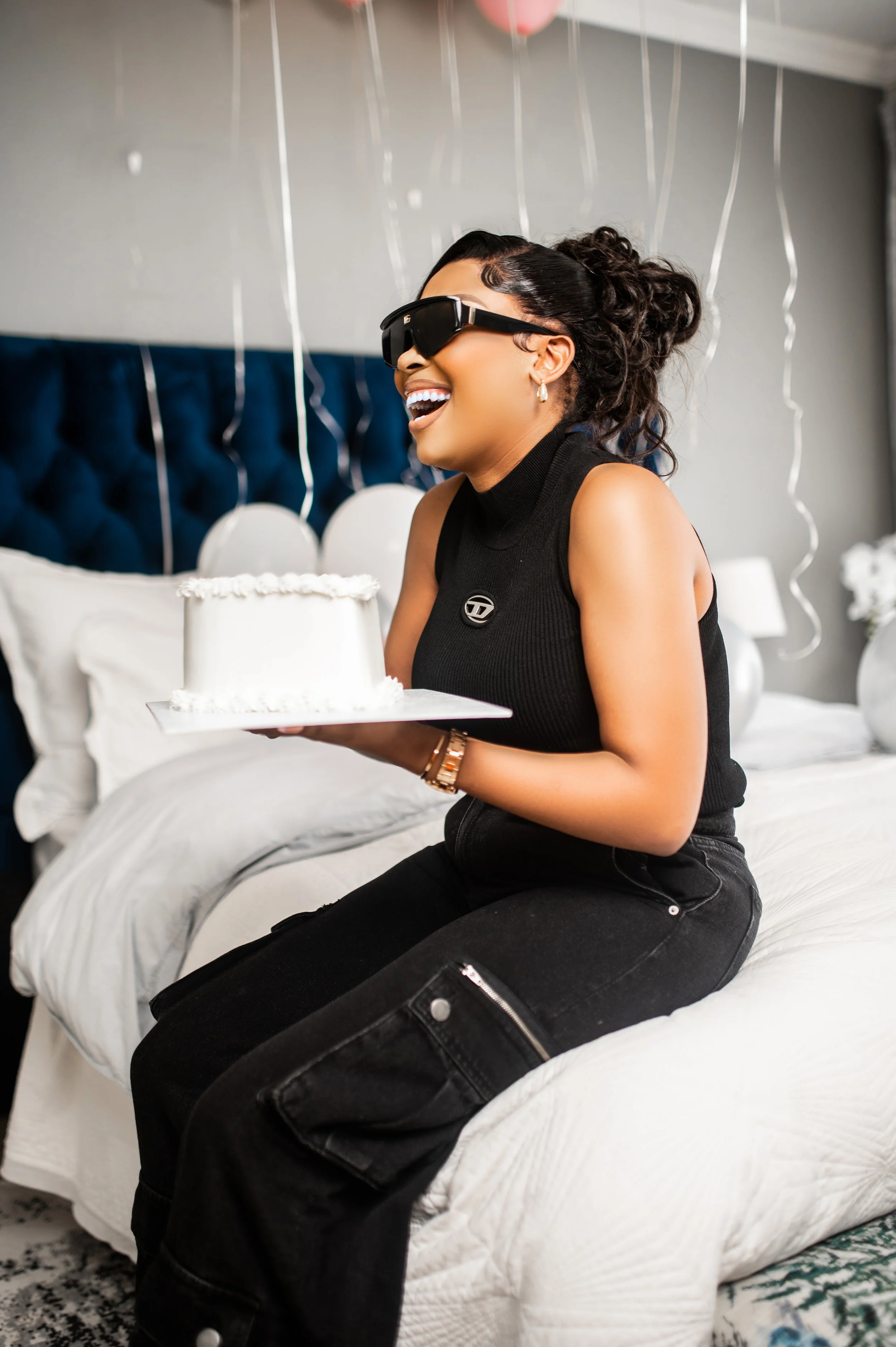 A woman sitting on a bed, smiling and holding a white frosted cake. She is wearing sunglasses and casual black clothing, surrounded by balloons and streamers, suggesting a celebration or party setting. Photographed in Melrose Arch, Johannesburg. 
