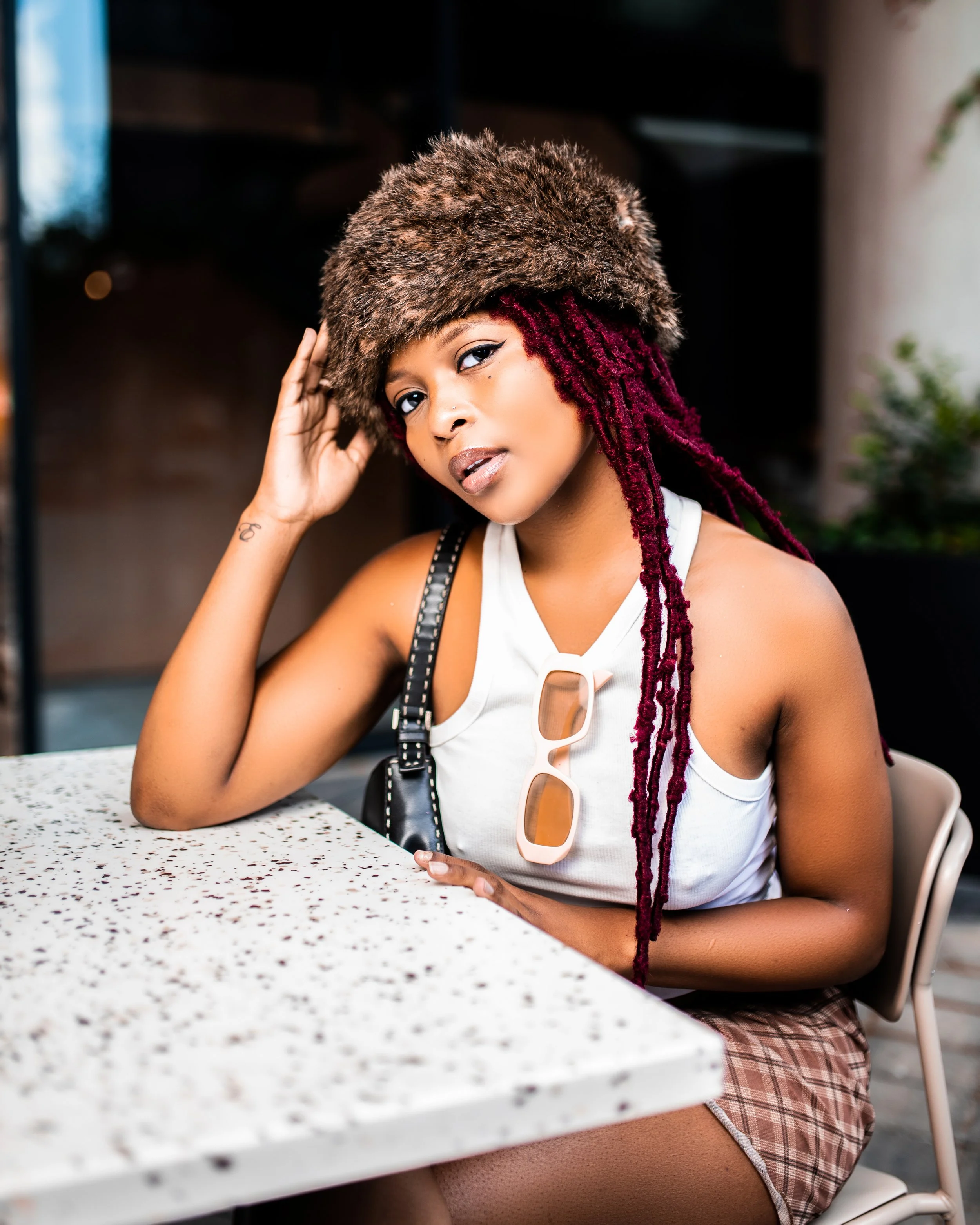 Person wearing a furry hat and white tank top sitting at a table. Photographed in Rosebank, South Africa.