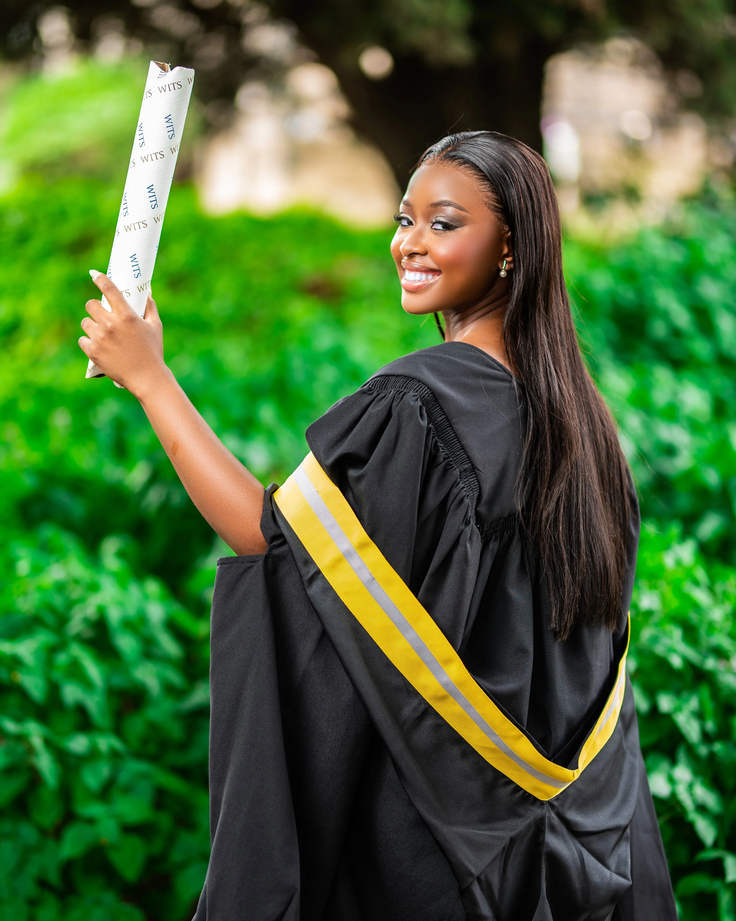 Graduate woman holding a diploma, wearing a black gown with yellow stripes, smiling in a green outdoor setting. Photographed at Wits University, Johannesburg. 
