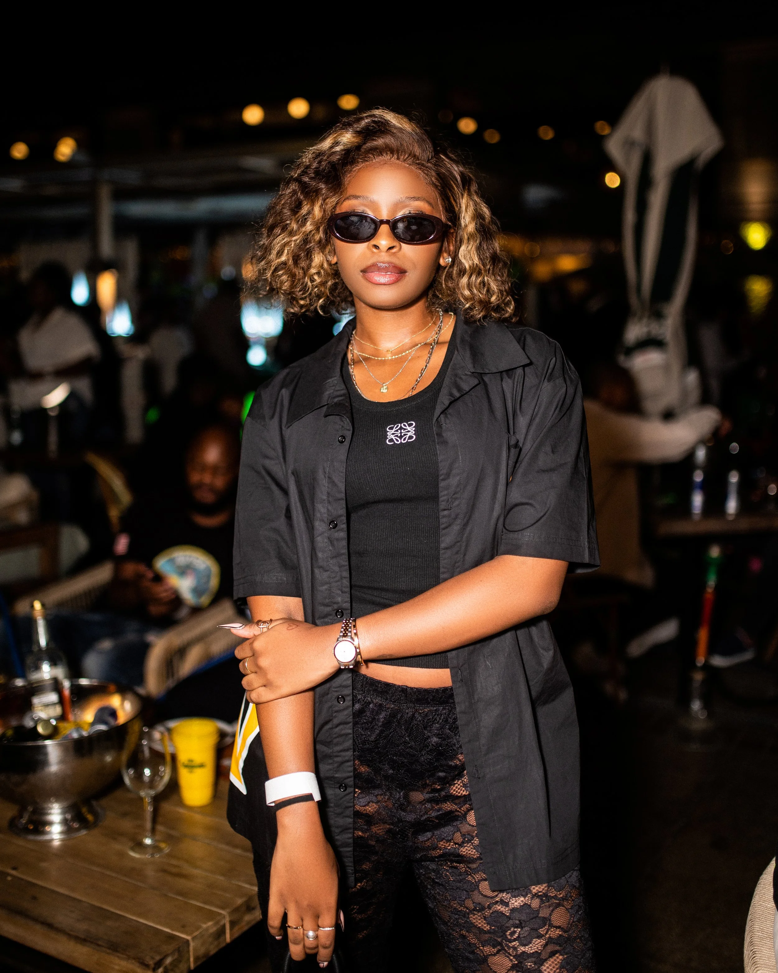 Person wearing black outfit and sunglasses at a dimly lit venue with tables and people in the background. Photographed at Rosebank, Johannesburg. 