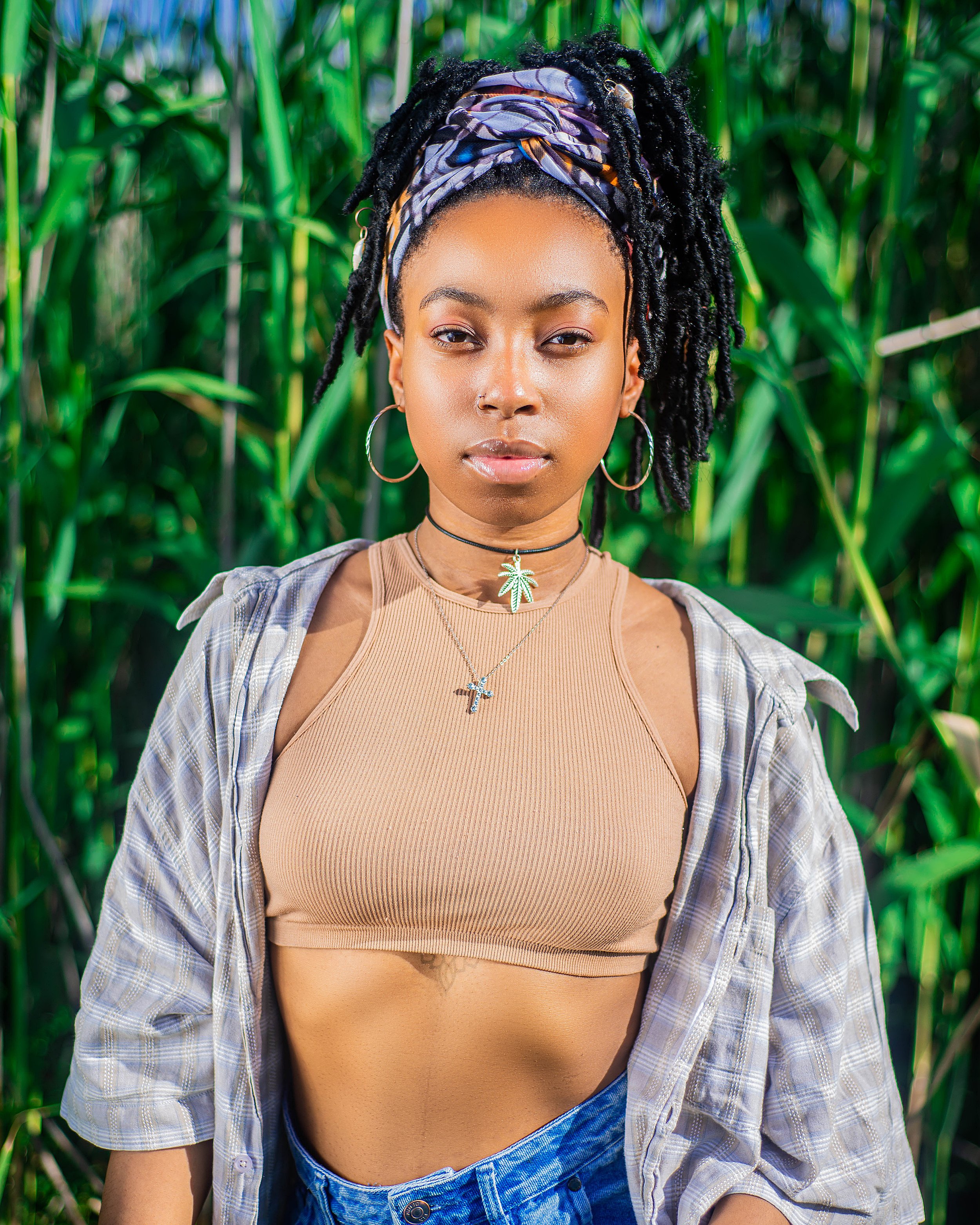 Young woman with headscarf and hoop earrings standing in front of tall plants, wearing a beige crop top, layered necklaces, plaid shirt, and jeans. Photographed in Auckland Park, Johannesburg.