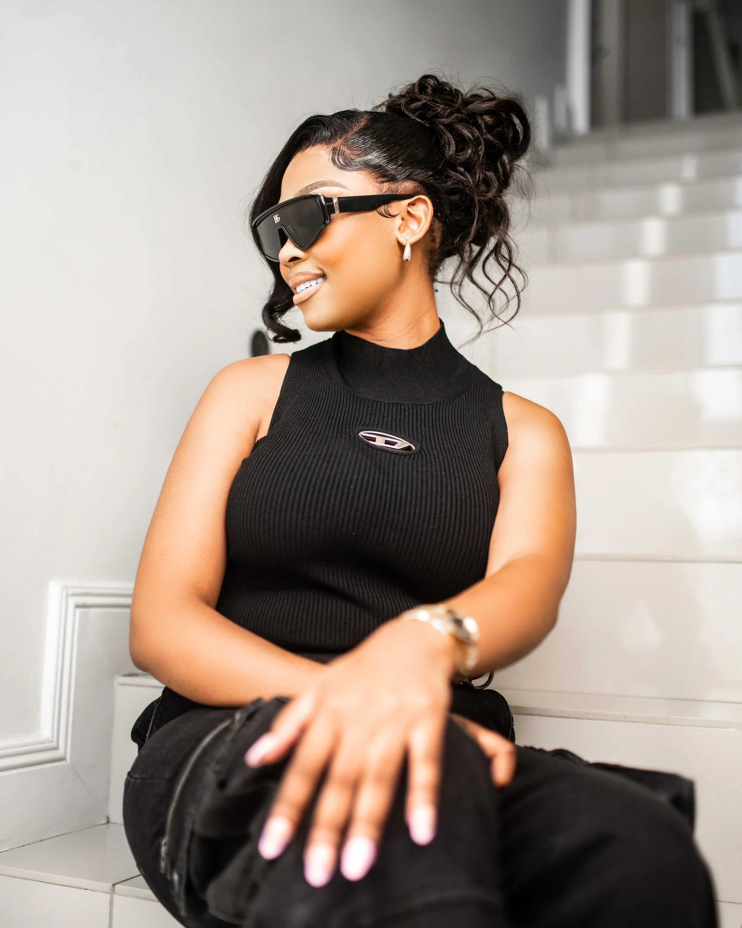Woman in black sleeveless top and sunglasses sitting on white stairs, smiling, with hands resting on knees. Photographed in Johannesburg, South Africa.