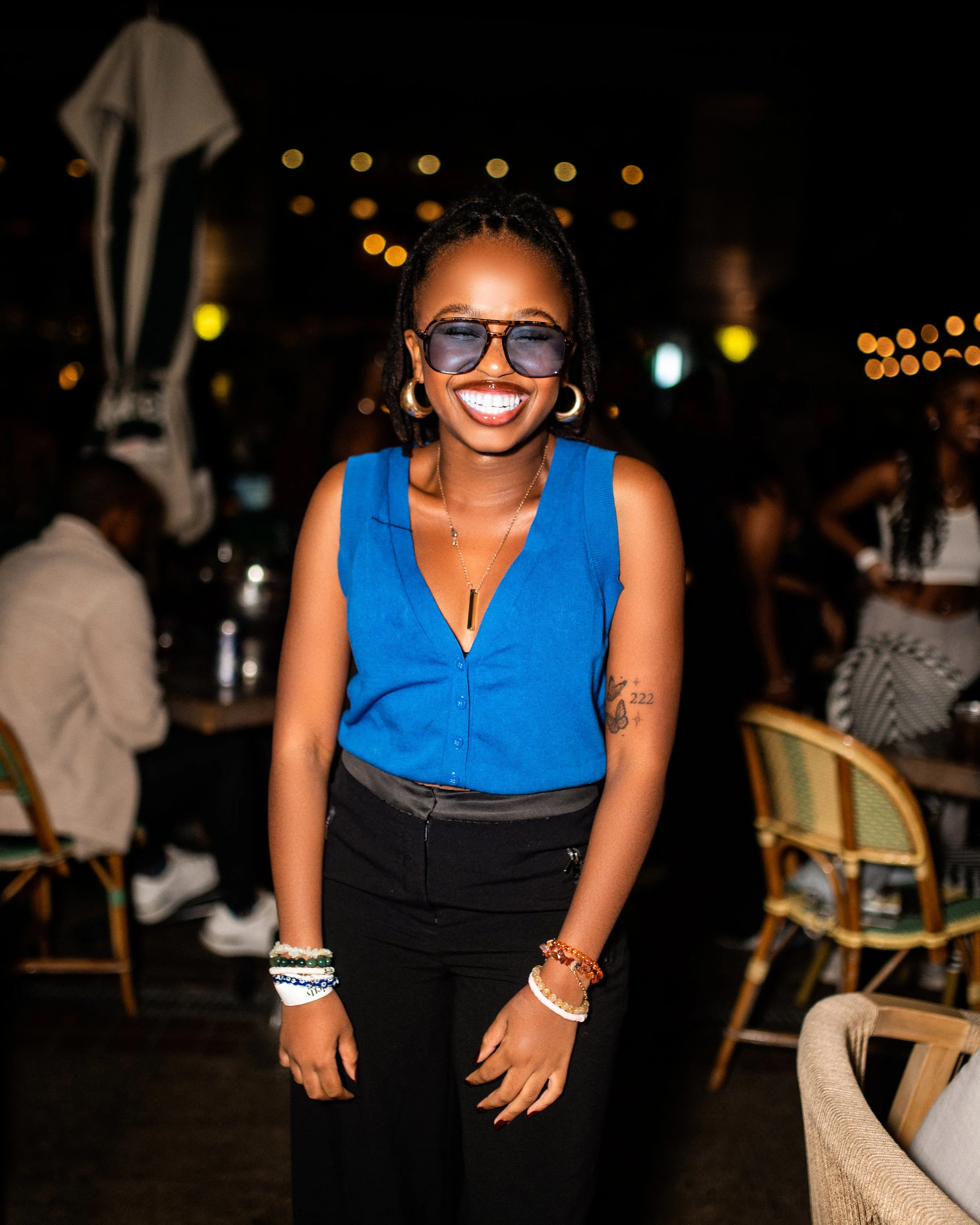 Smiling woman in blue top and sunglasses at night, with blurred background of people and lights.  Photographed in Rosebank, Johannesburg, South Africa.