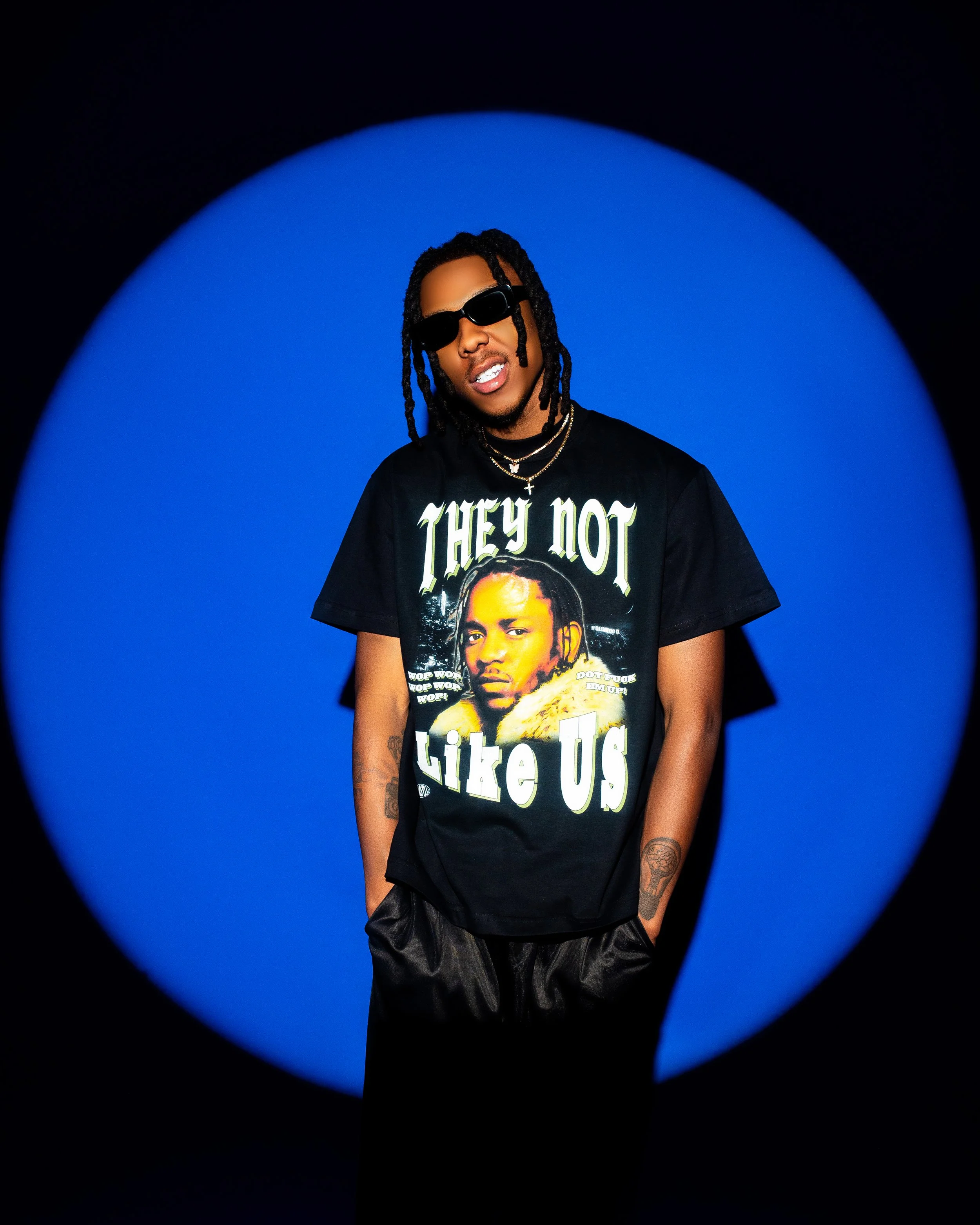 A person wearing sunglasses and a black T-shirt with the phrase 'They Not Like Us' printed on it stands in front of a blue backdrop. Photographed in Bryanston, Johannesburg.