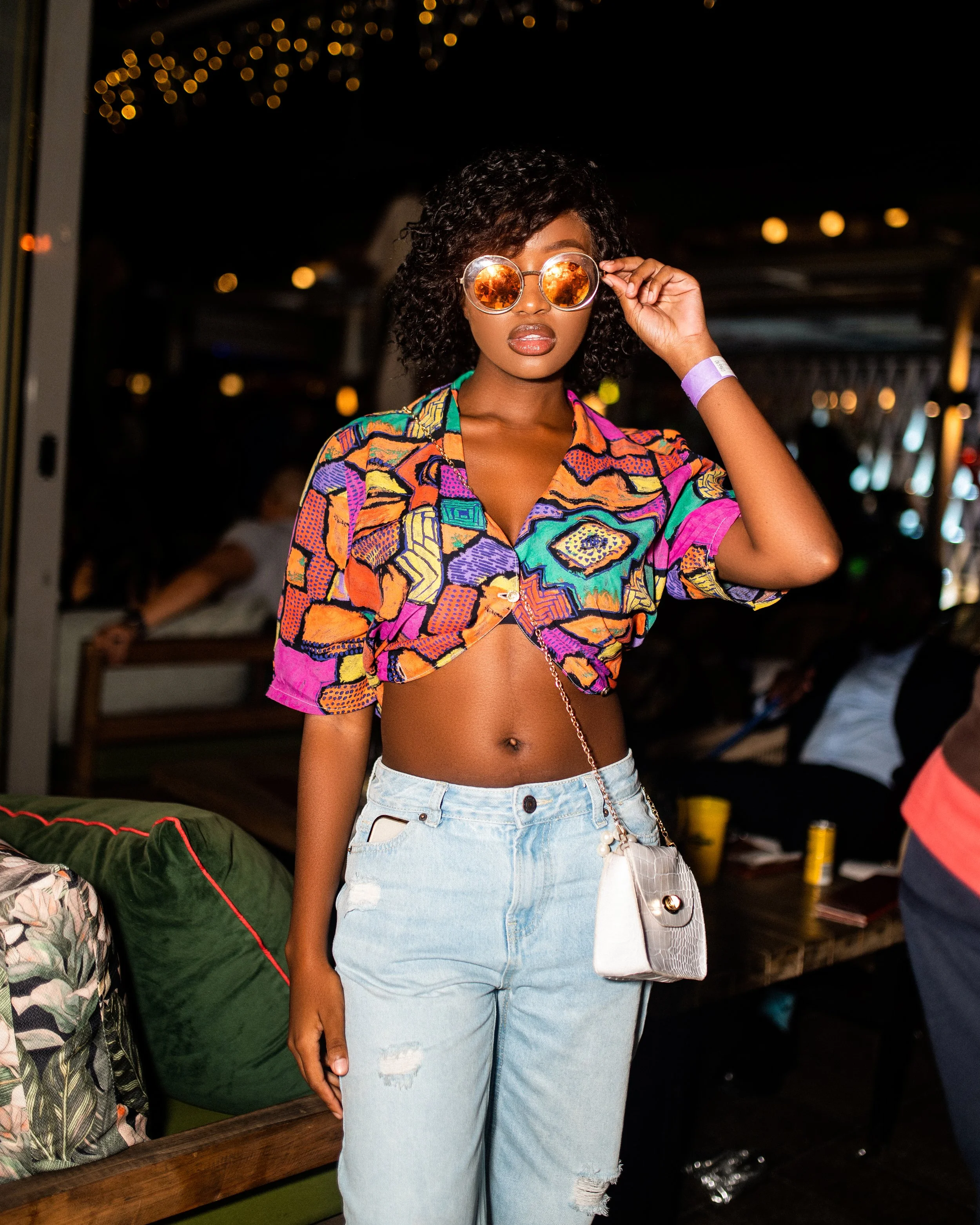 Woman wearing colorful patterned top, jeans, and orange sunglasses at night. Photographed in Rosebank, South Africa.