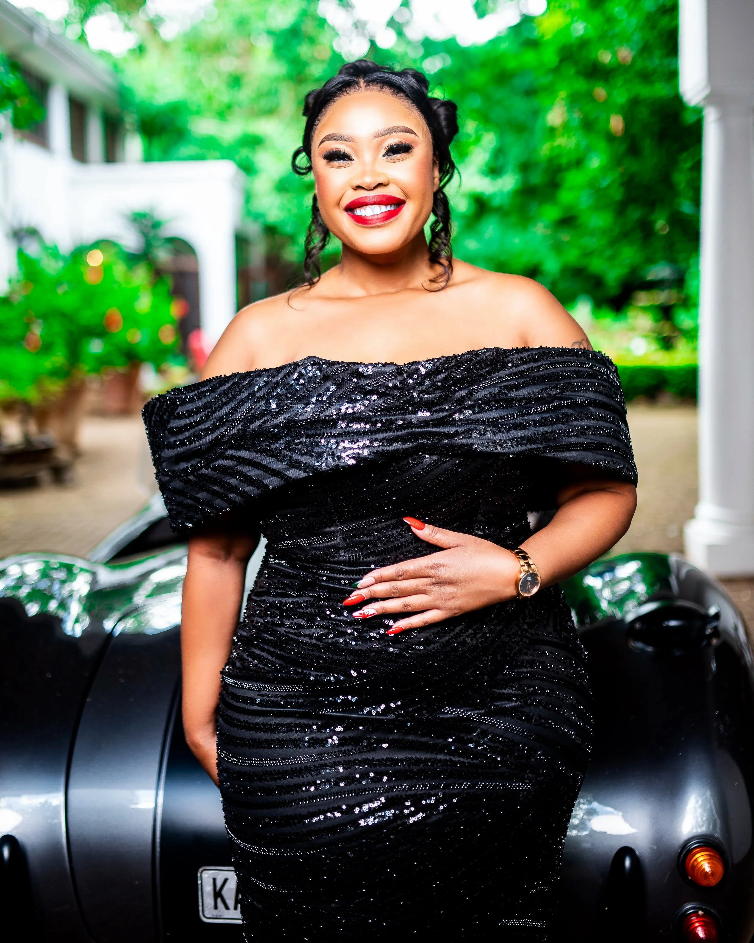 Woman in off-shoulder black sequin dress standing in front of a car, with lush greenery in the background. Photographed in Johannesburg, South Africa.