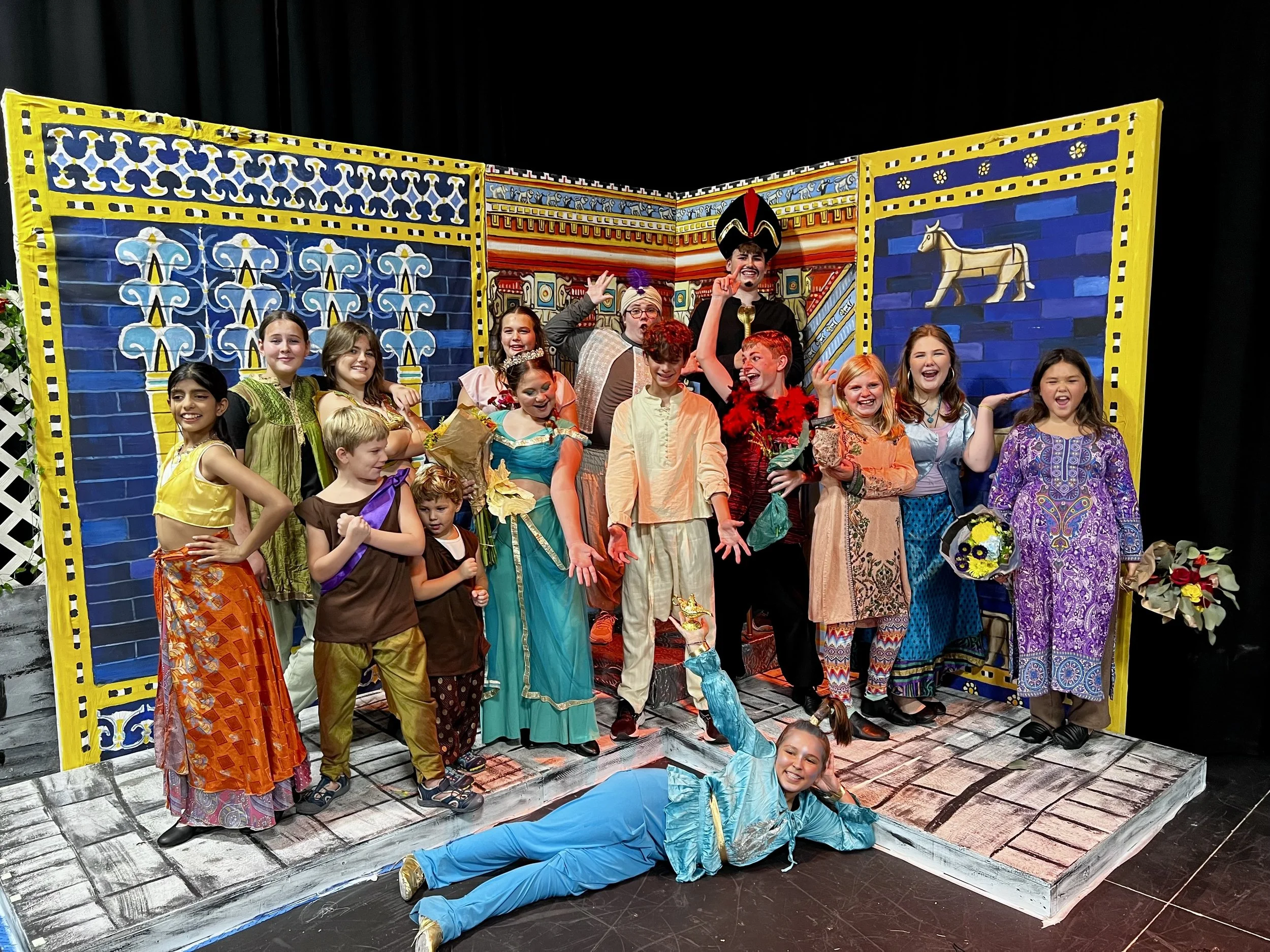 Children in colorful costumes on stage with a vibrant, theatrical backdrop resembling a royal palace scene. A girl in blue lying on the stage floor, smiling, with other kids standing behind her, some holding props and flowers.