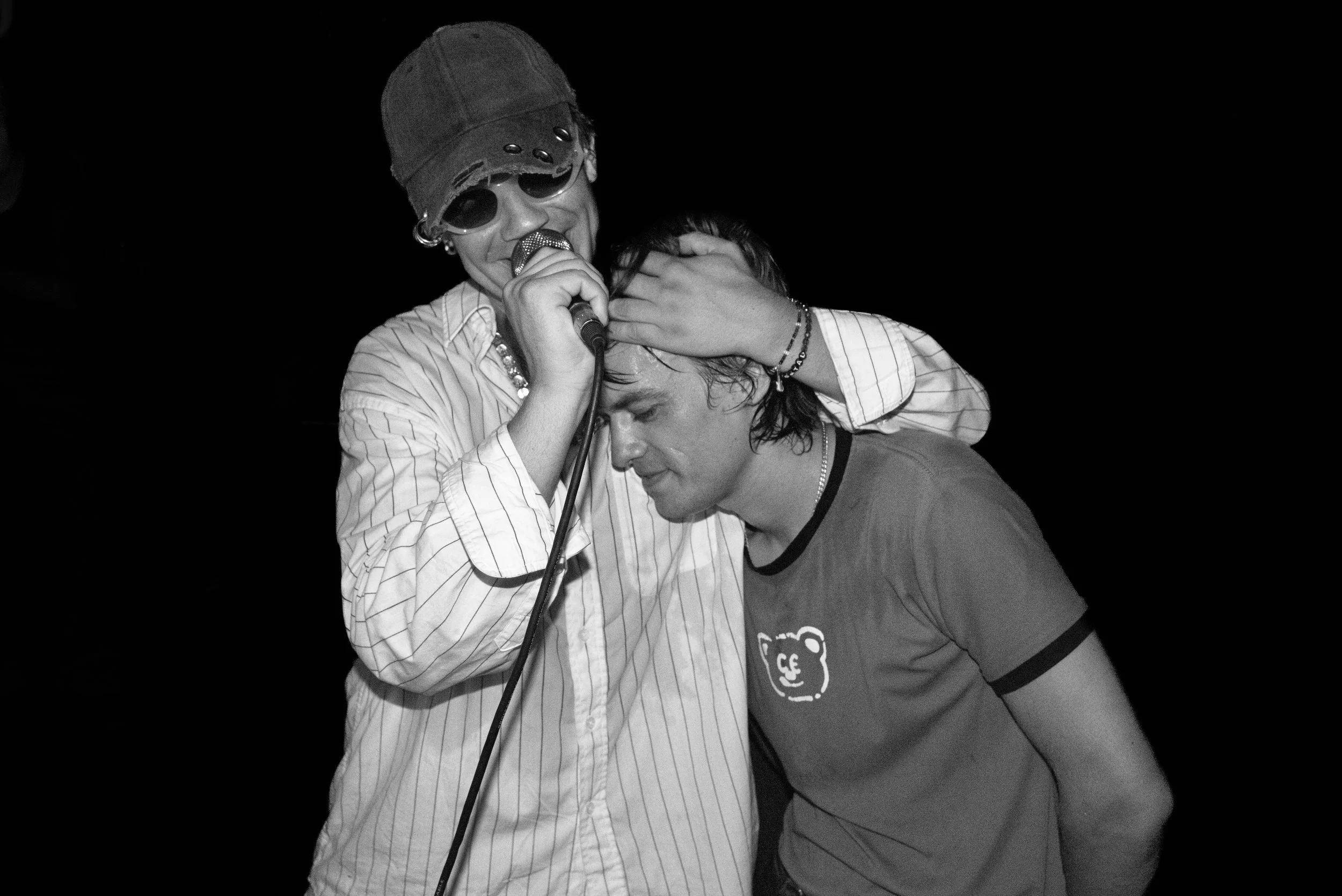 Two young men, one wearing a cap and sunglasses, holding a microphone with his eyes closed, and the other with wet hair, leaning into the first with eyes closed, against a dark background.