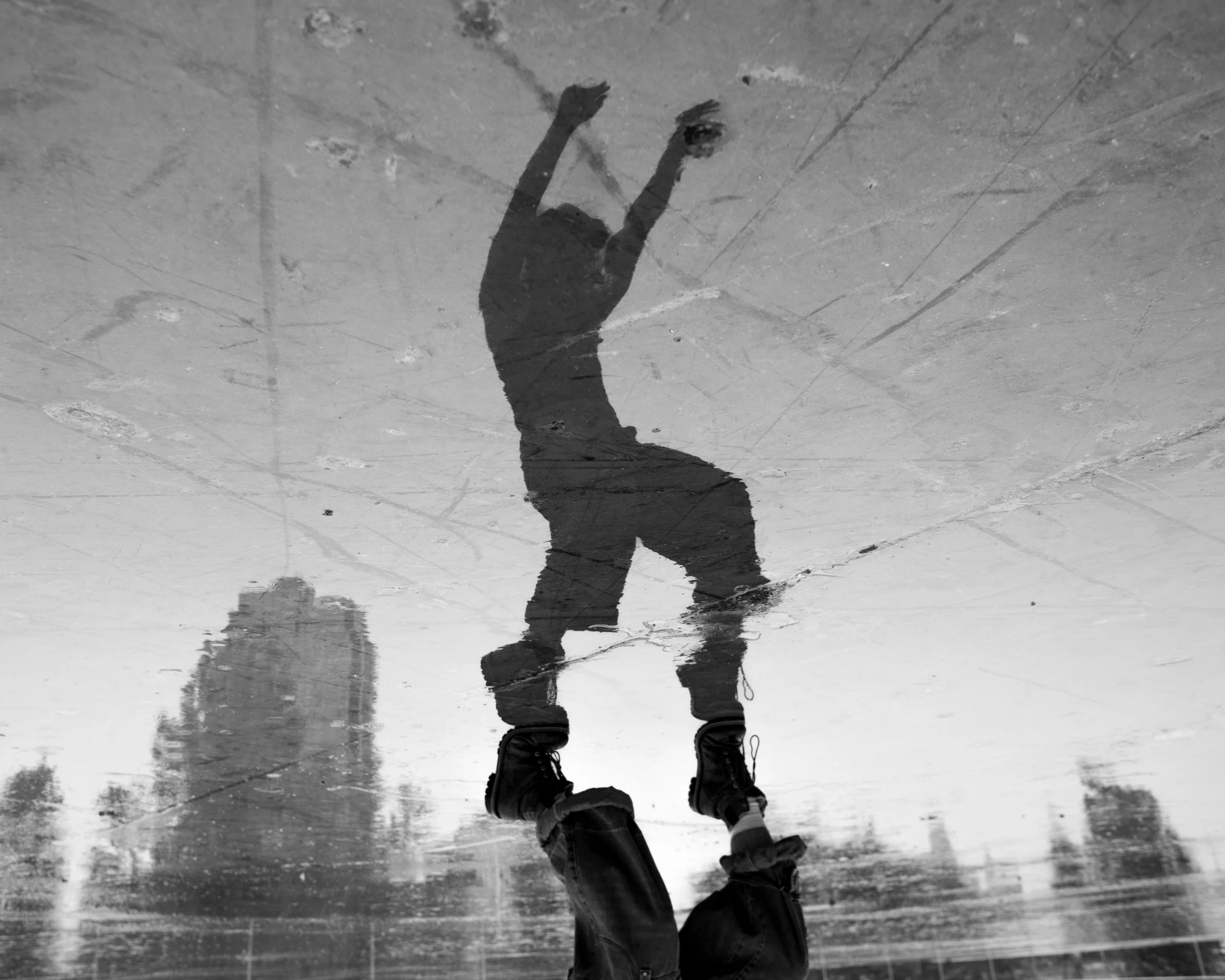 Reflection of a person skating on a wet pavement, with trees and buildings visible in the background.