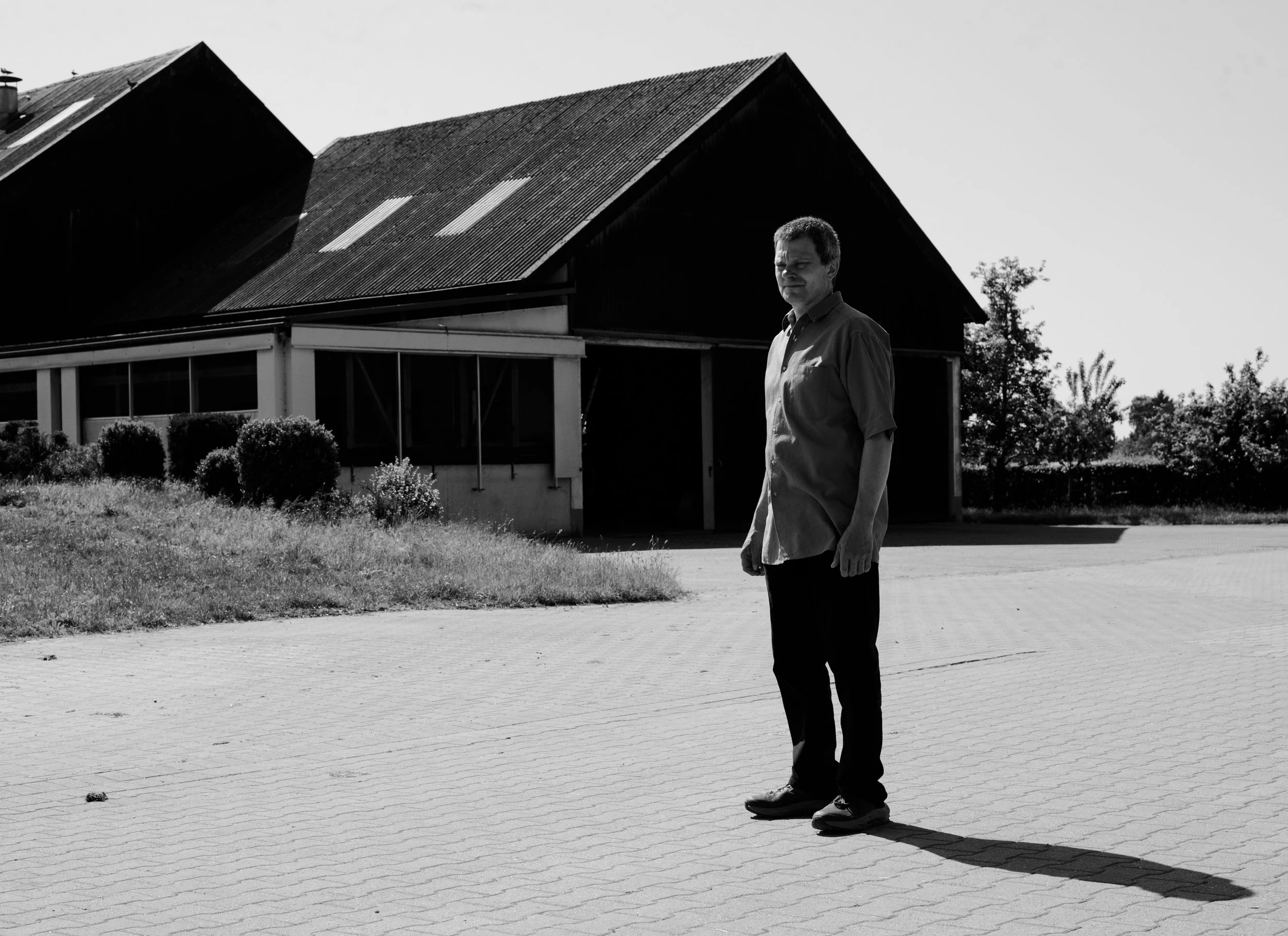 A man stands on a paved area outside a barn, with his shadow cast long on the ground in bright sunlight. The barn has a metal roof and surrounded by shrubs and trees.