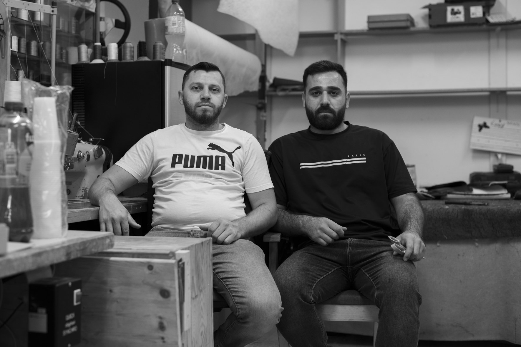 Two men sitting on a bench in a workshop or store, surrounded by tools and materials, looking at the camera.