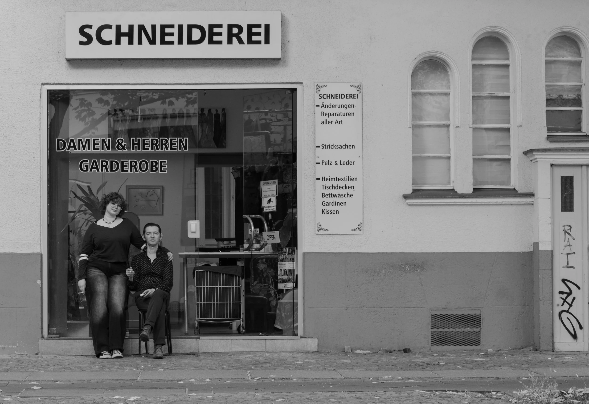 A black and white photo of a clothing store with large window display, showing two women in front, one standing and one seated, with the store sign reading 'Schneiderei' and a list of services and items in German.