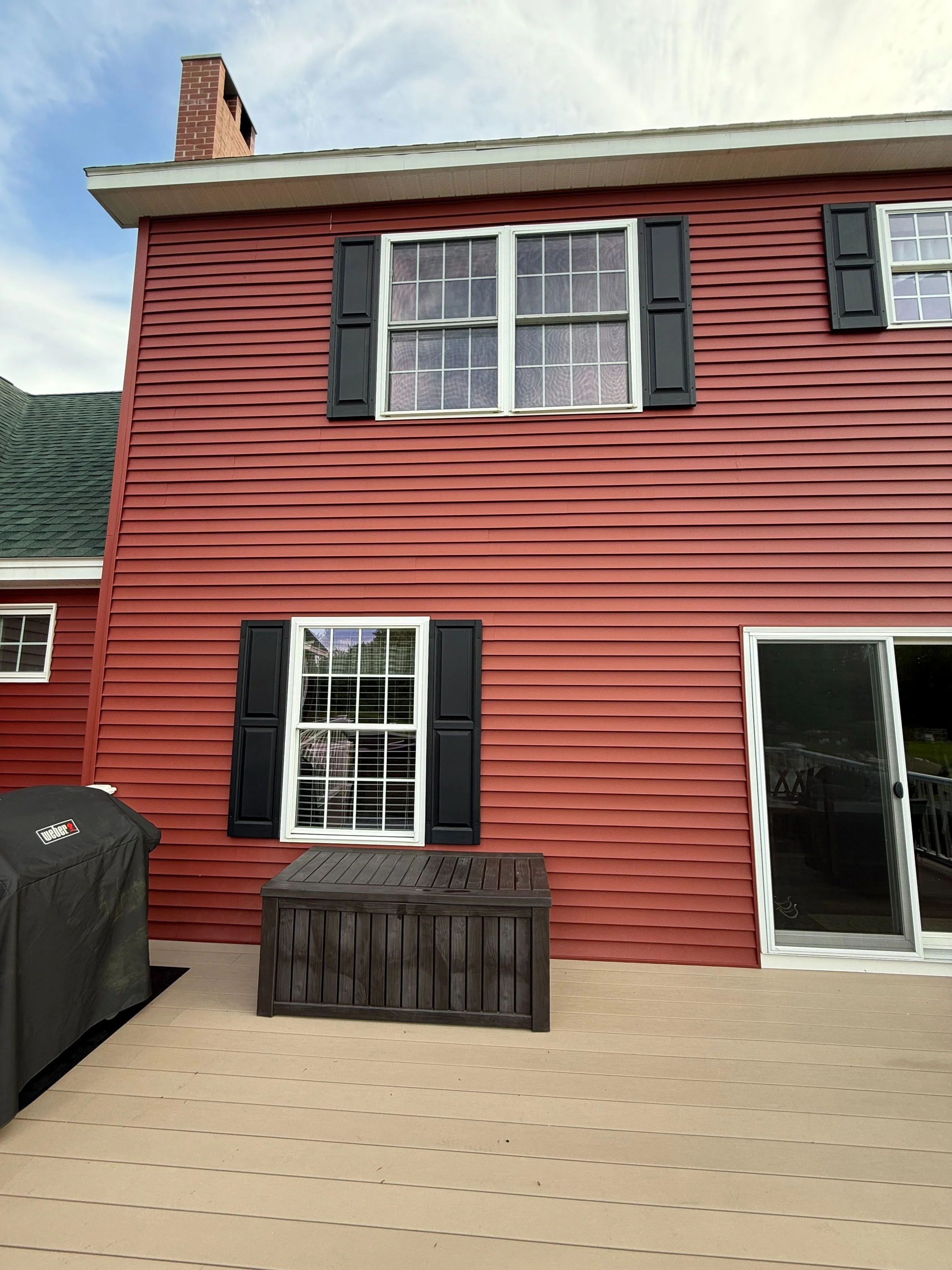 Red house with black shutters, attached to a deck with a black grill and a wooden storage box, three windows and a sliding glass door, overcast sky.