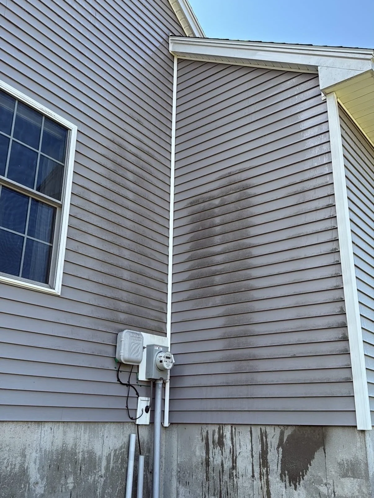 Exterior wall of a house with beige siding, black soot stains, a window with white trim, and an electric meter box attached at the bottom
