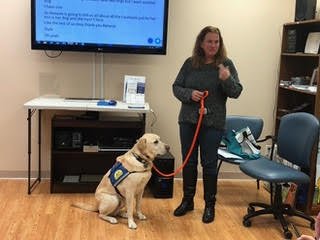 A woman standing beside a service dog in a room with a screen displaying text. The dog is wearing a harness, and the woman is holding its leash. They appear to be part of a presentation or training session. There are chairs and a table with electroni