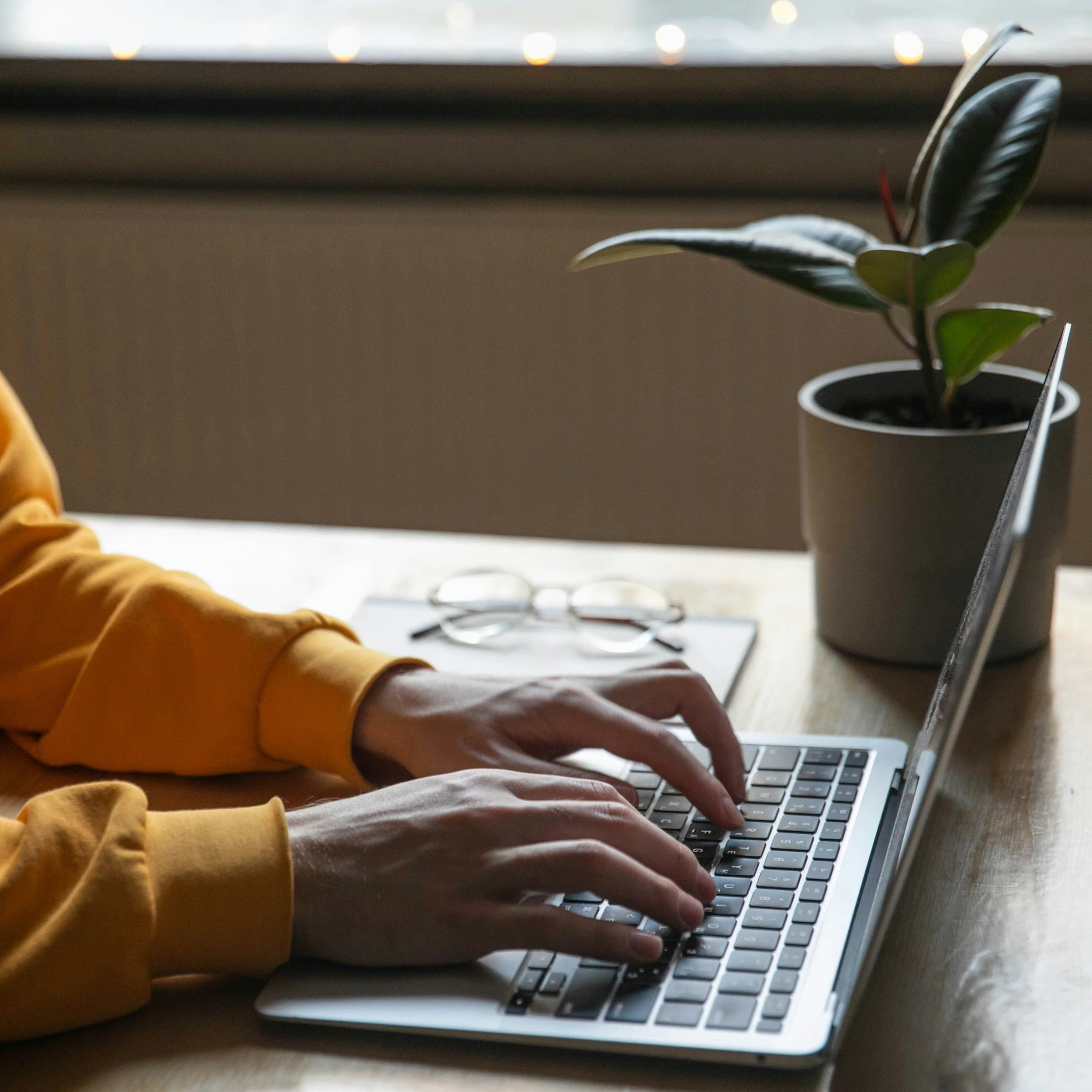 Person wearing a yellow sweatshirt typing on a silver laptop at a wooden desk, with a pair of eyeglasses, a notebook, and a potted plant nearby.