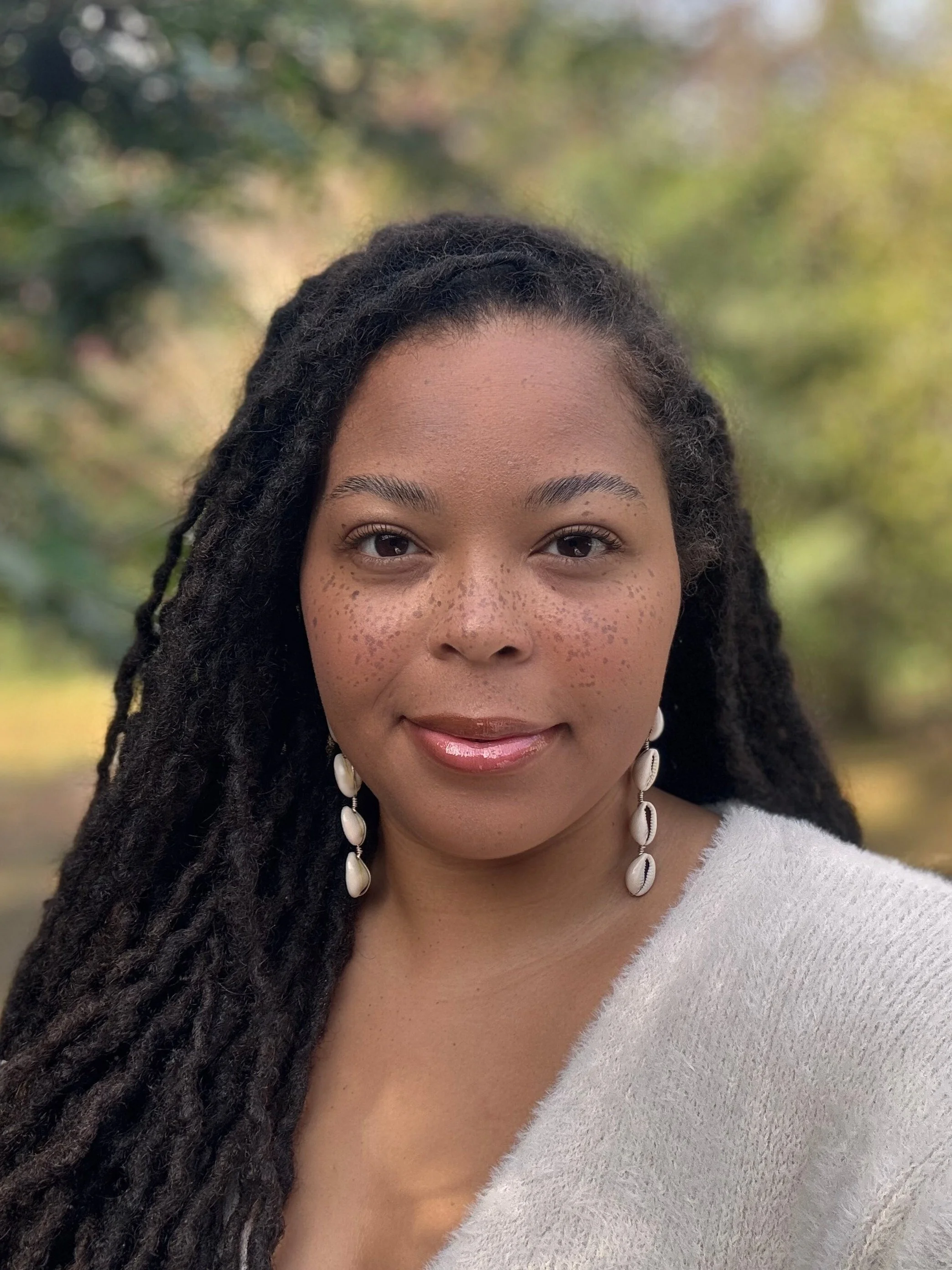 Close-up of a woman with long, dark dreadlocks, wearing white shell earrings and a white sweater, outdoors with blurred green foliage in the background.