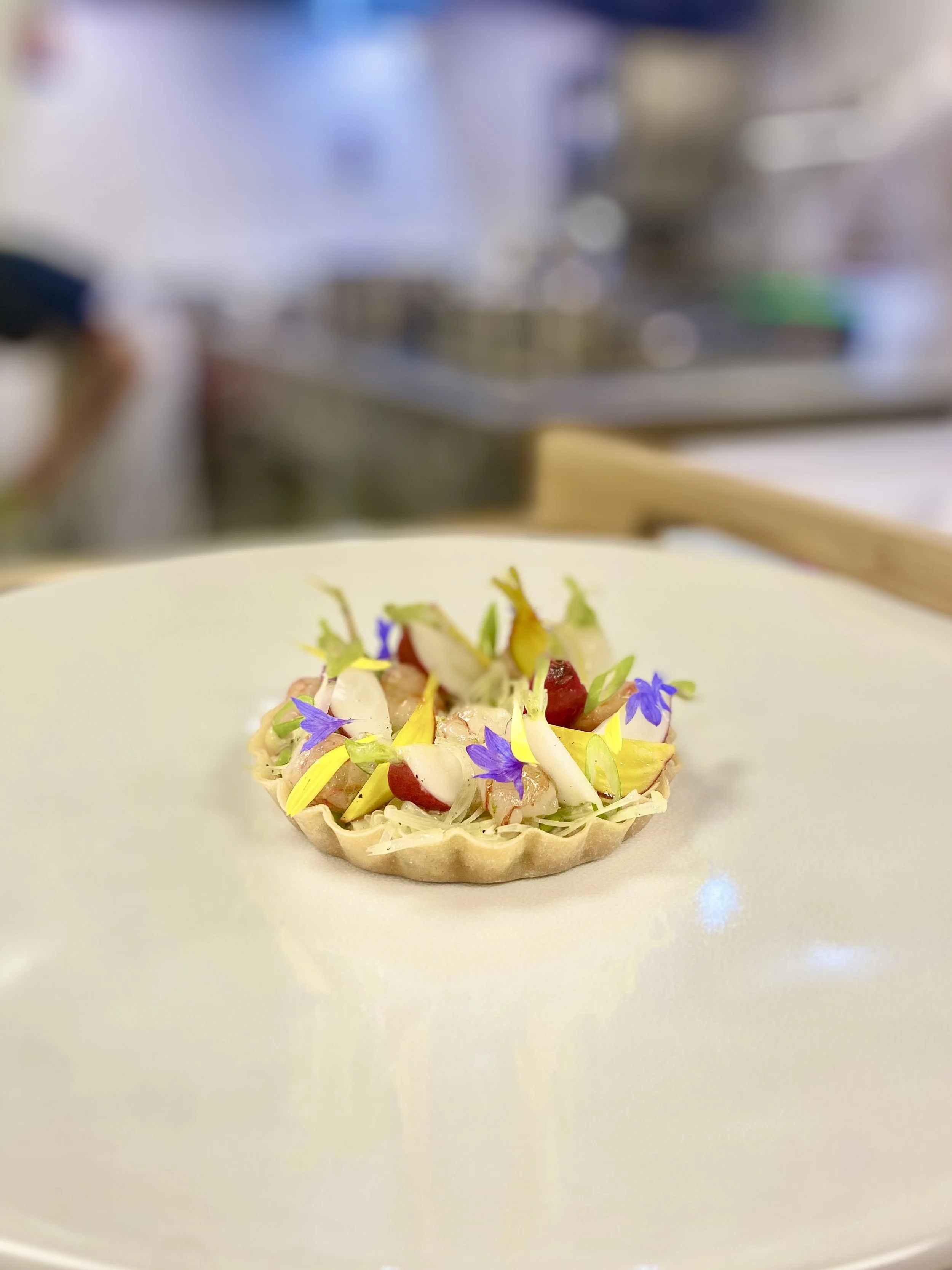 A small, colorful edible art dish on a white plate in a kitchen setting, garnished with edible flowers and vegetables.