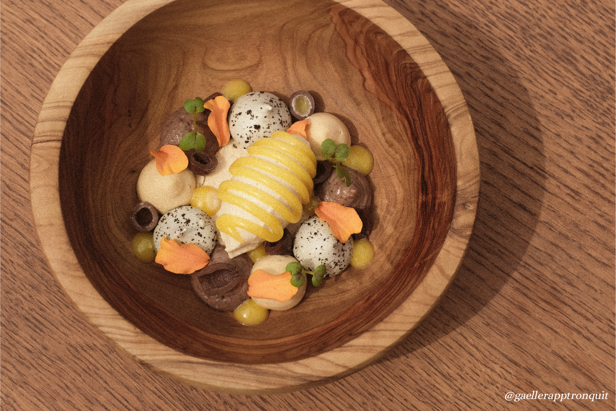 A dessert in a wooden bowl, including a swirl of yellow sauce, small chocolates, green microgreens, and orange edible flowers.