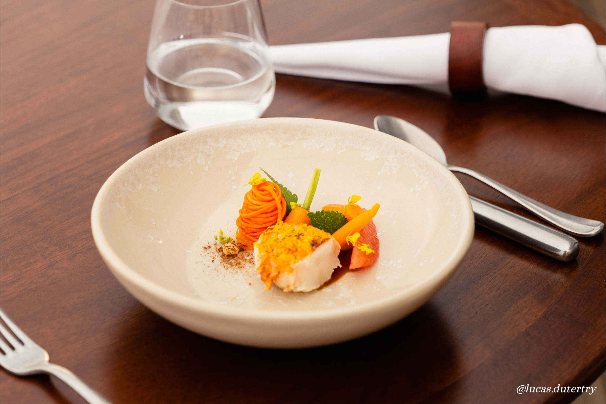 A plated modern gourmet dish featuring a piece of white fish with crispy topping, served with orange carrot puree, garnished with green herbs and yellow flower petals, positioned on a cream-colored bowl on a wooden table with silverware, a glass of w