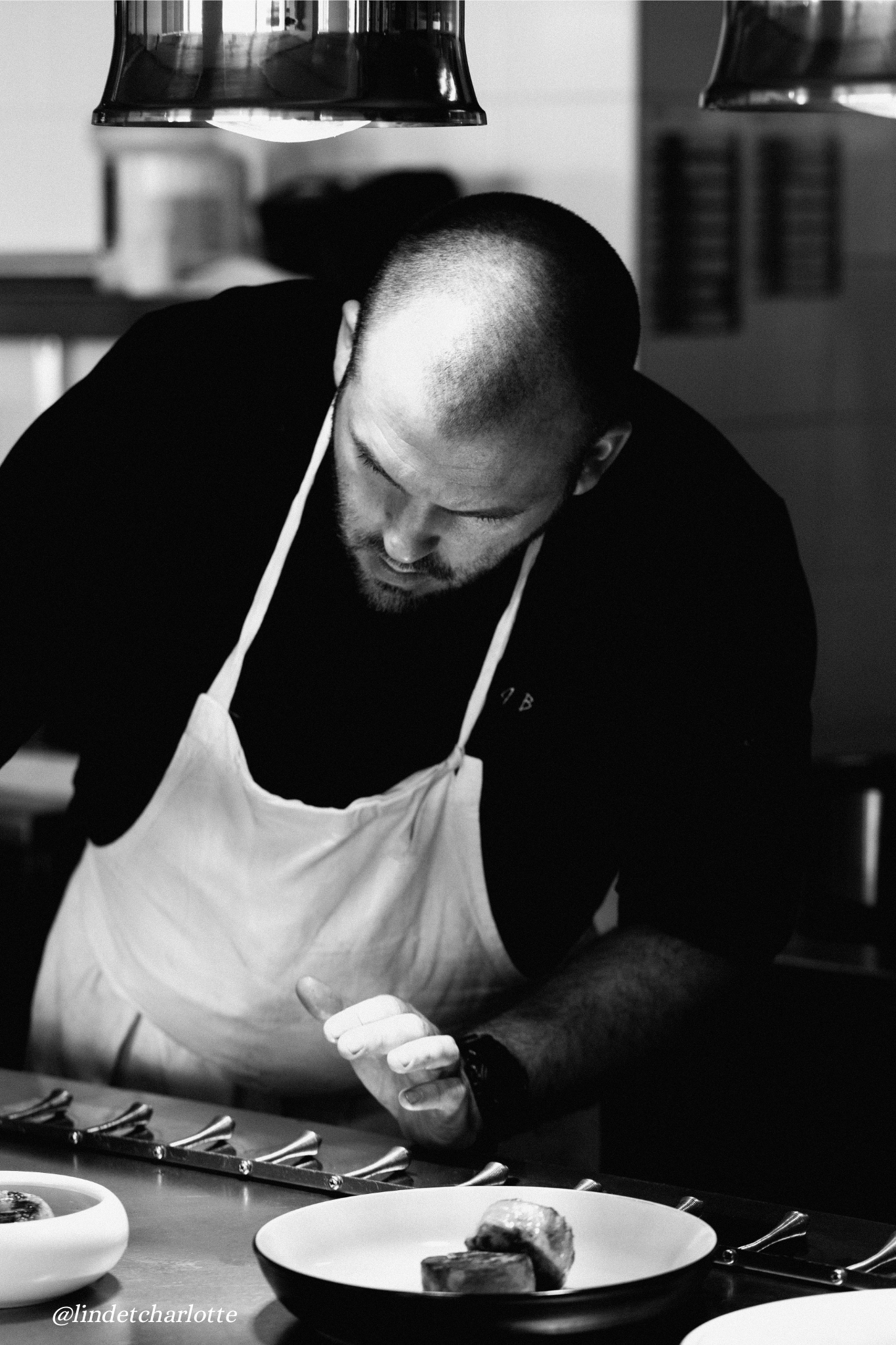 A man in a black shirt and white apron is preparing food in a kitchen. He is looking down at the food, focusing on his work. There are dishes with food on the counter in front of him. The photo is in black and white.