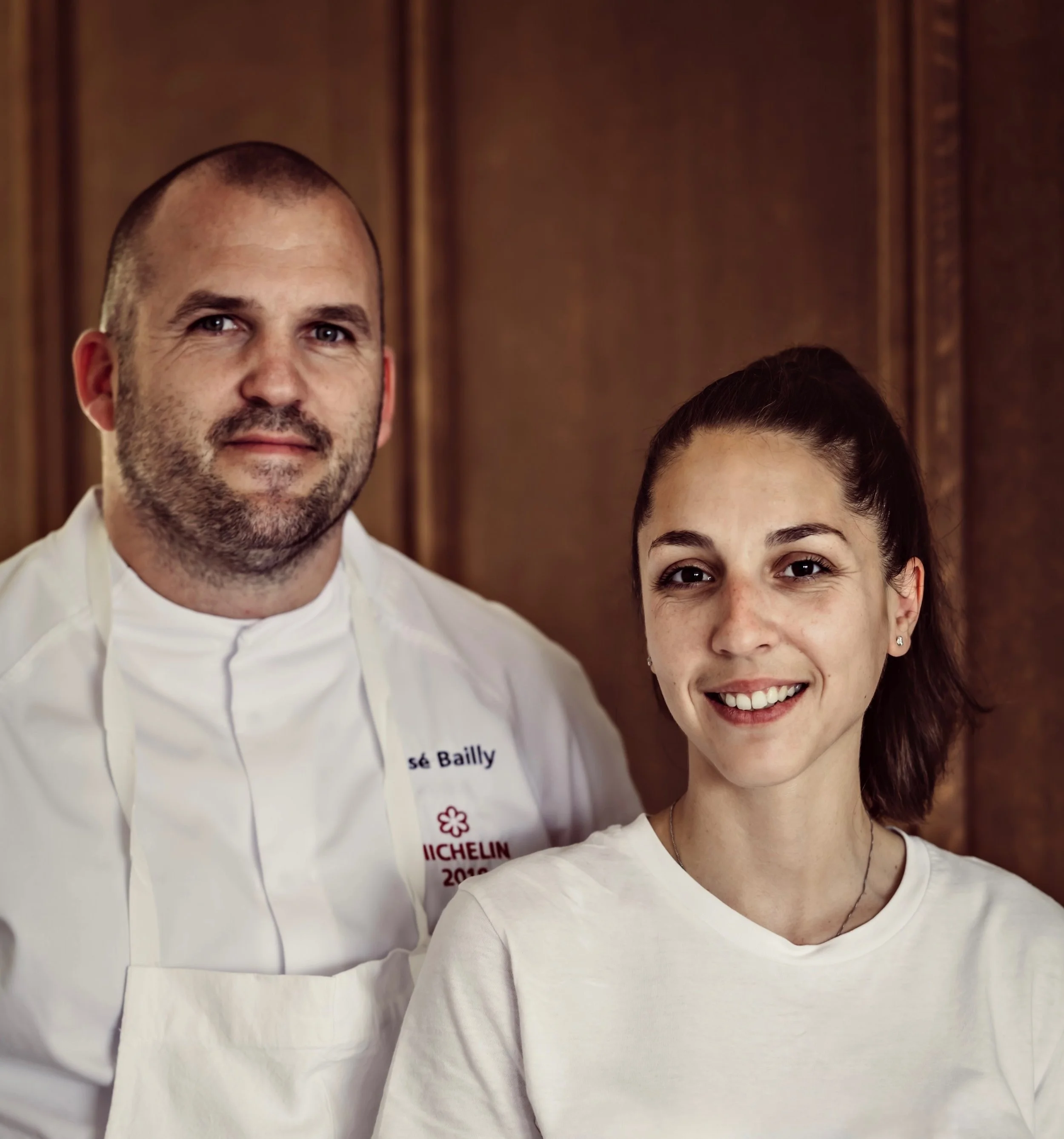 Close-up of a man with a shaved head and beard wearing a white chef's coat and a woman with dark hair and earrings wearing a white shirt, both smiling, against a wood-paneled background.