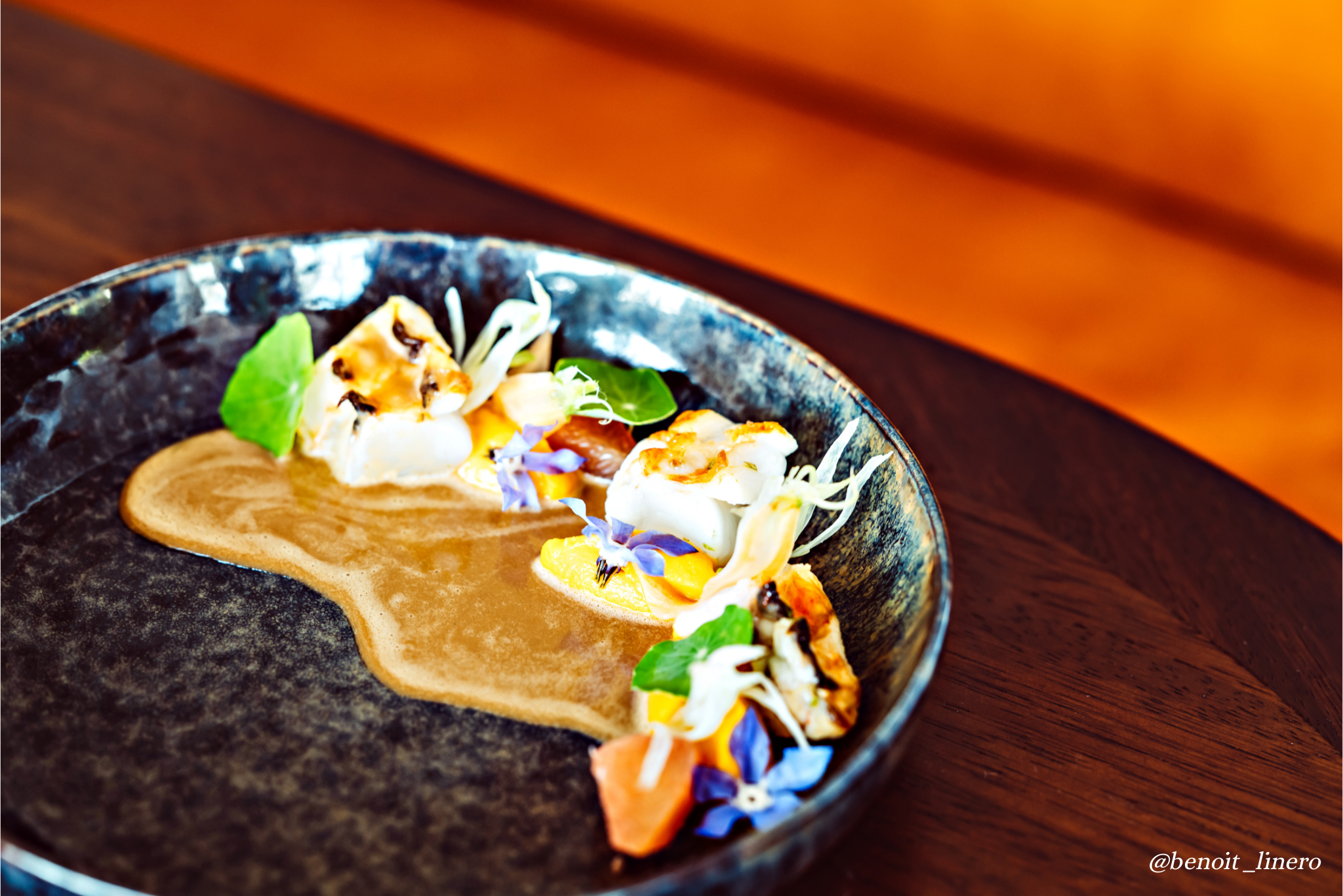 Elegant plate with seared scallops, colorful edible flowers, greens, and a brown sauce on a black textured plate placed on a wooden surface.