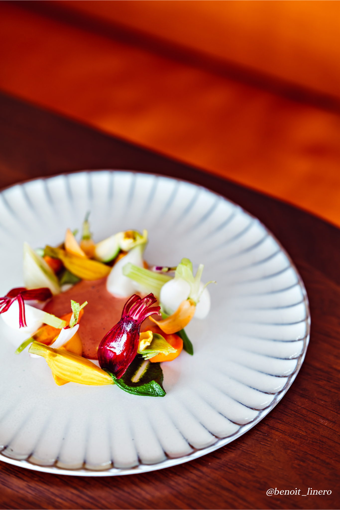A plated gourmet dish with colorful edible flowers and vegetables on a white, textured plate set on a wooden table.