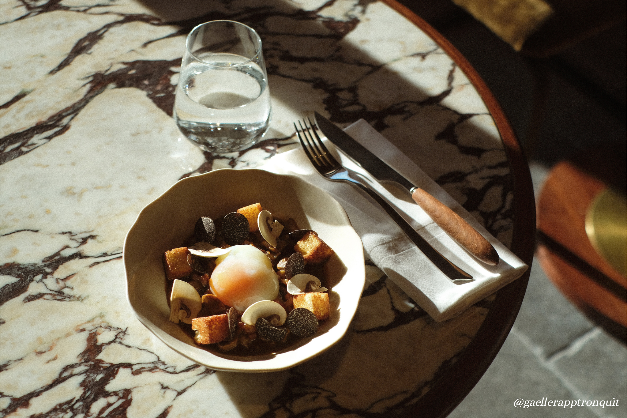 A marble table with a glass of water, a bowl of dessert with a soft-boiled egg, and a napkin with silverware.