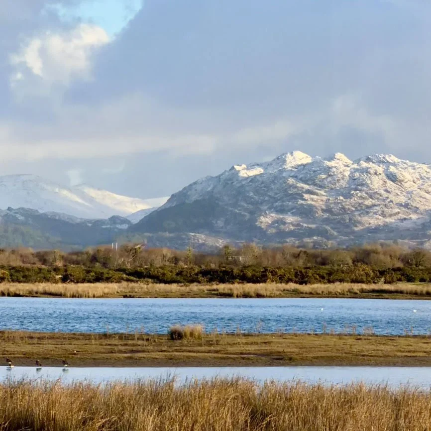 Landscape view of a lake with turquoise water surrounded by mountains and grassy hills under a cloudy sky.