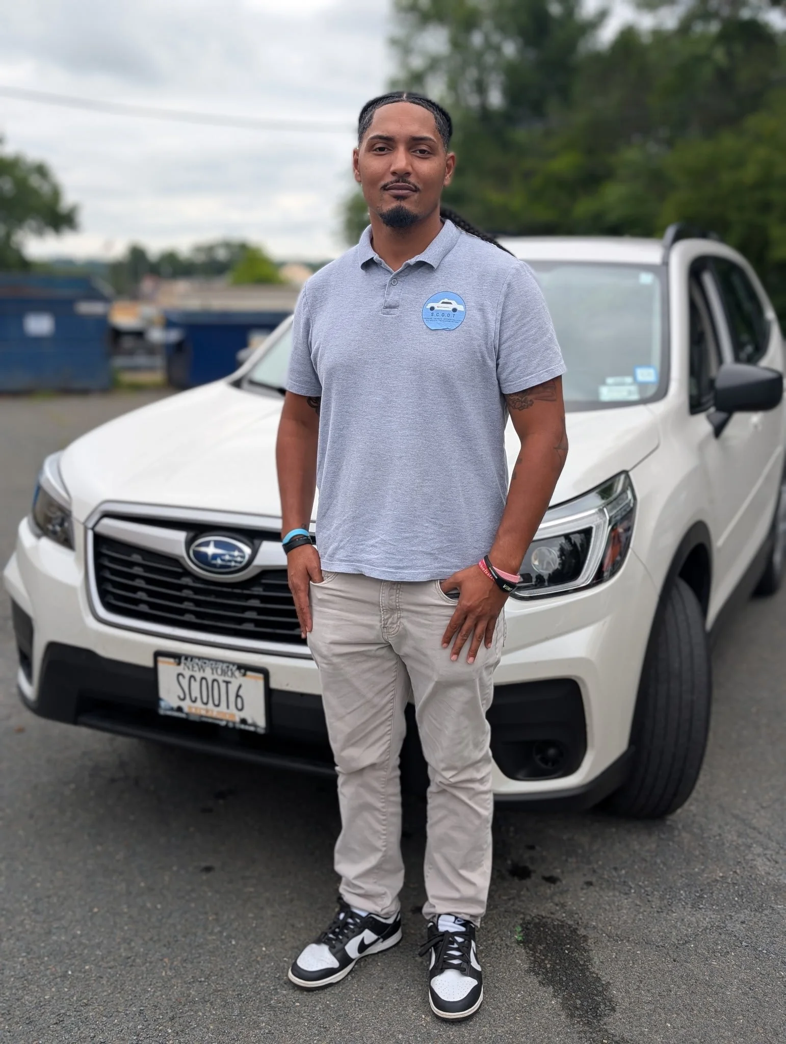 Young man with braided hair and a goatee standing outdoors next to a black car, wearing a light-colored sweatshirt, black vest, and a necklace with a cross, in front of a brick building.