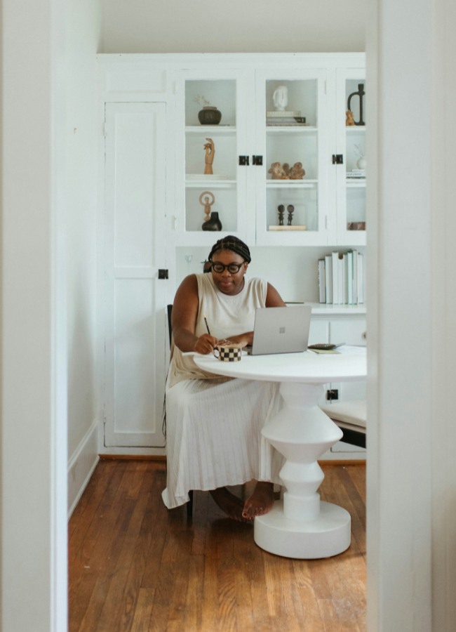 Photo of woman making notes next to her laptop, while sat at a dining room table.