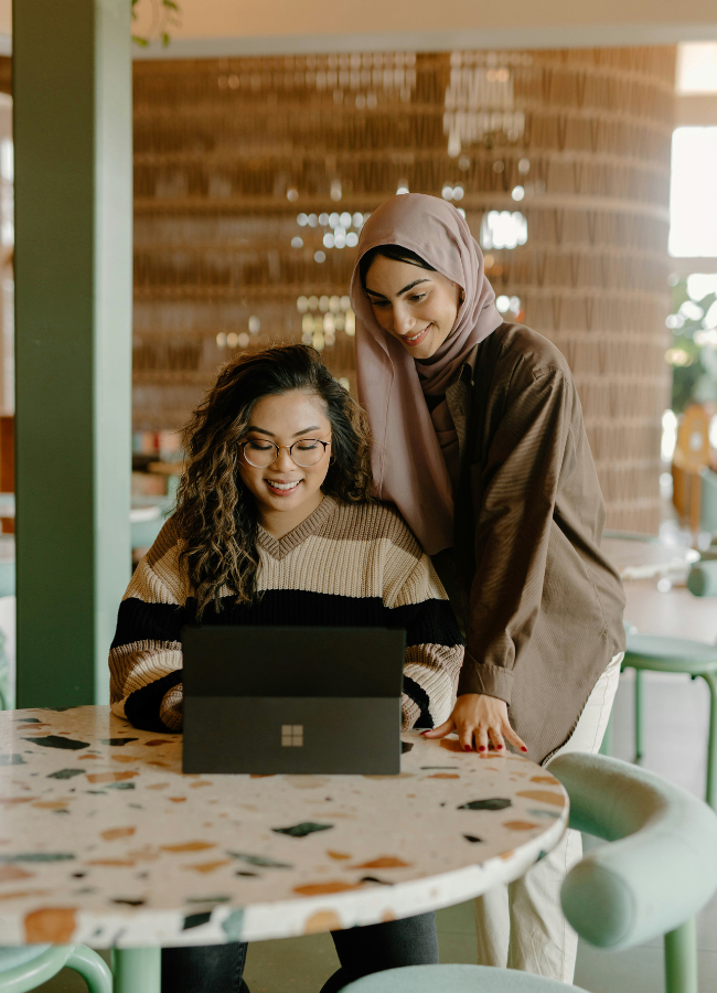 Photo of two woman looking at a laptop while sitting inside a cafe